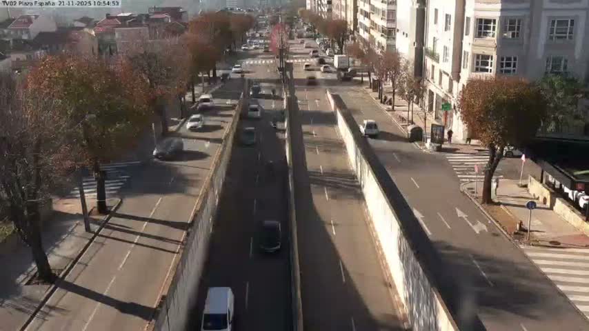 An overhead view on a clear, sunny day shows a multi-lane urban street with a central underpass, bustling with vehicles and flanked by buildings and trees with autumn foliage.