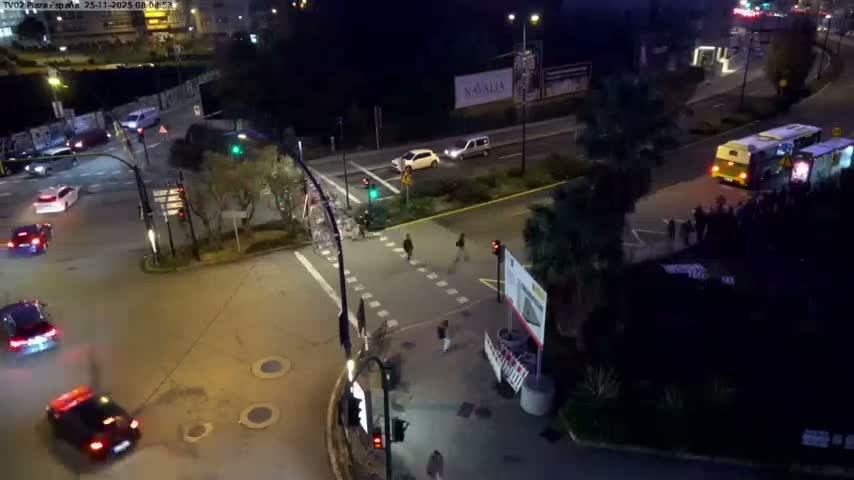 A busy urban intersection is depicted at night with vehicles on the roads, pedestrians crossing and waiting, and streetlights illuminating the scene under clear weather.