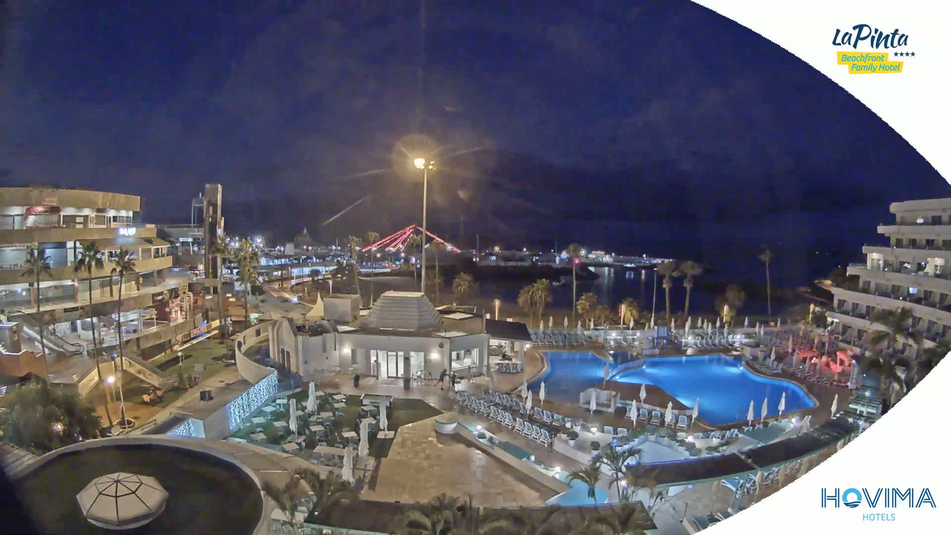 A nighttime aerial view of a resort complex with a swimming pool, illuminated buildings, and a harbor in the background under a clear, dark sky.