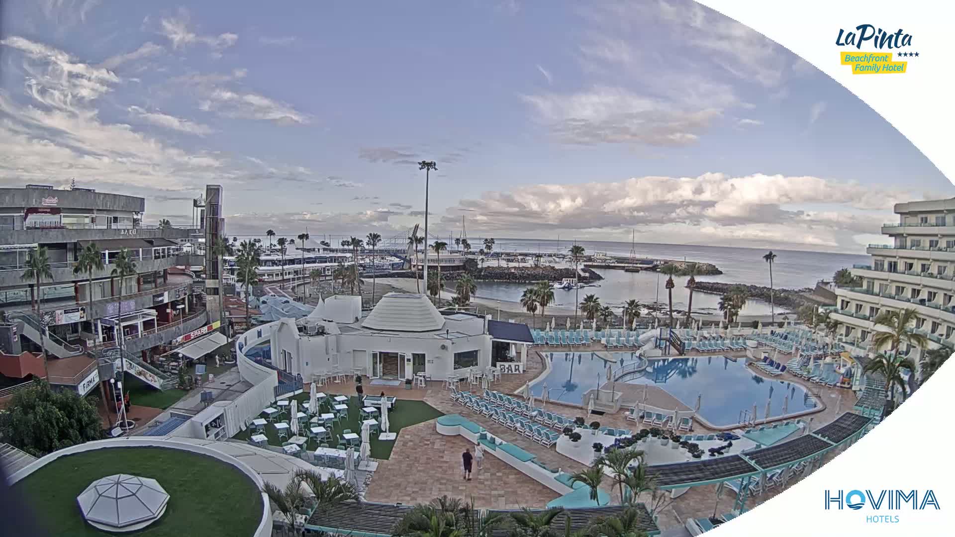 A resort pool area with a bar and surrounding buildings overlooks a calm harbor under a partly cloudy sky.