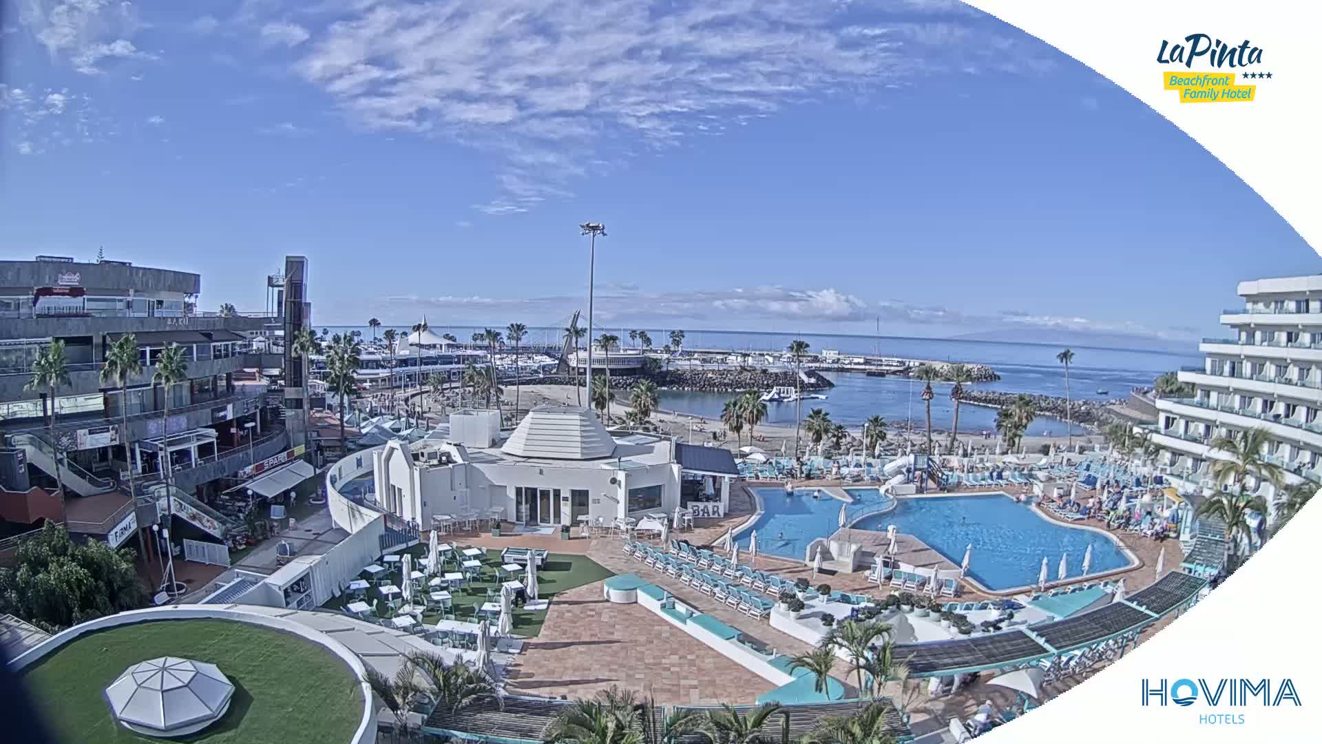 A sunny day overlooks a resort with a swimming pool, beach, and marina, under a partly cloudy sky.