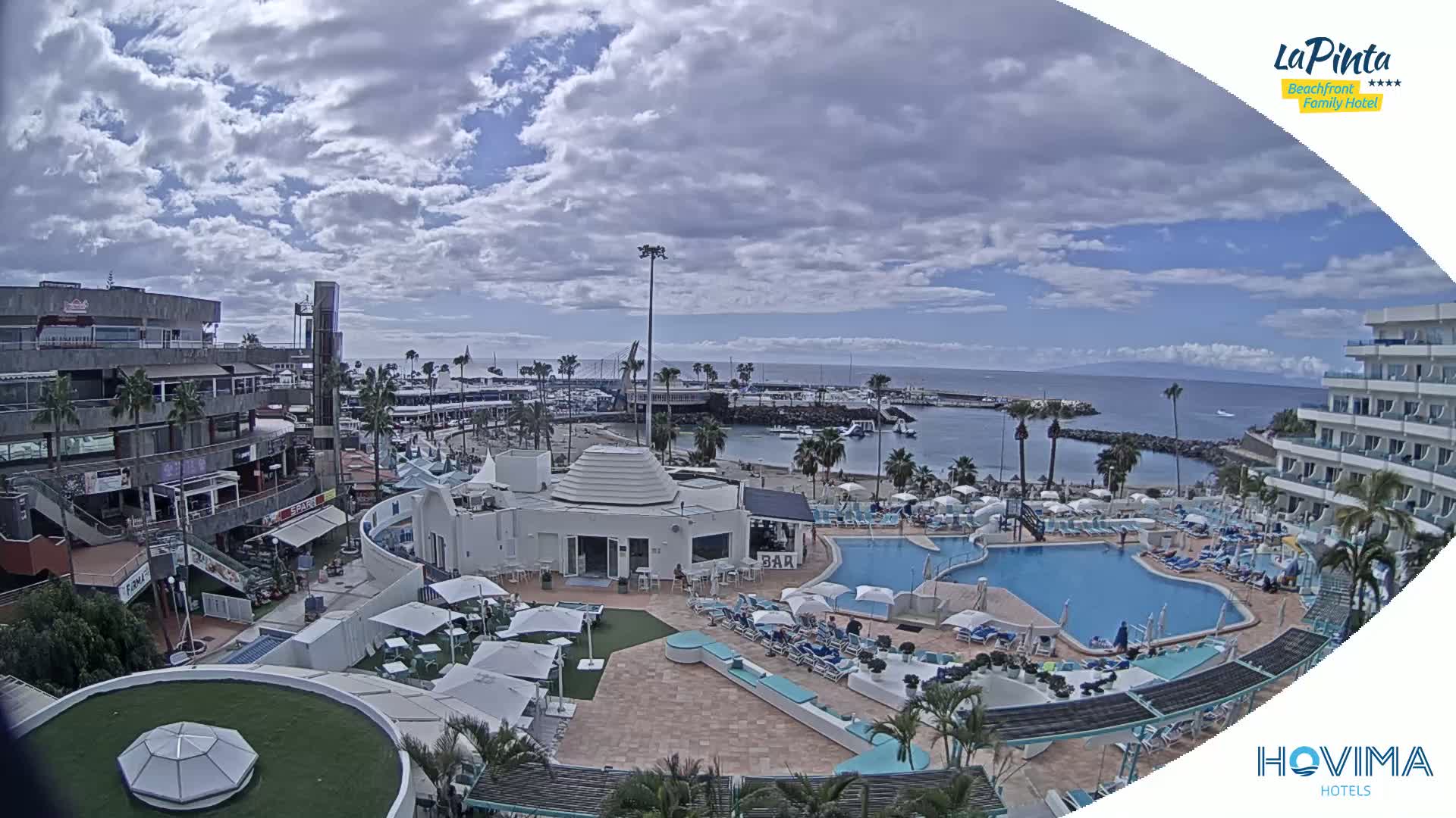 A resort pool area with a bar and surrounding buildings overlooks a partly cloudy harbor with boats and a calm ocean.