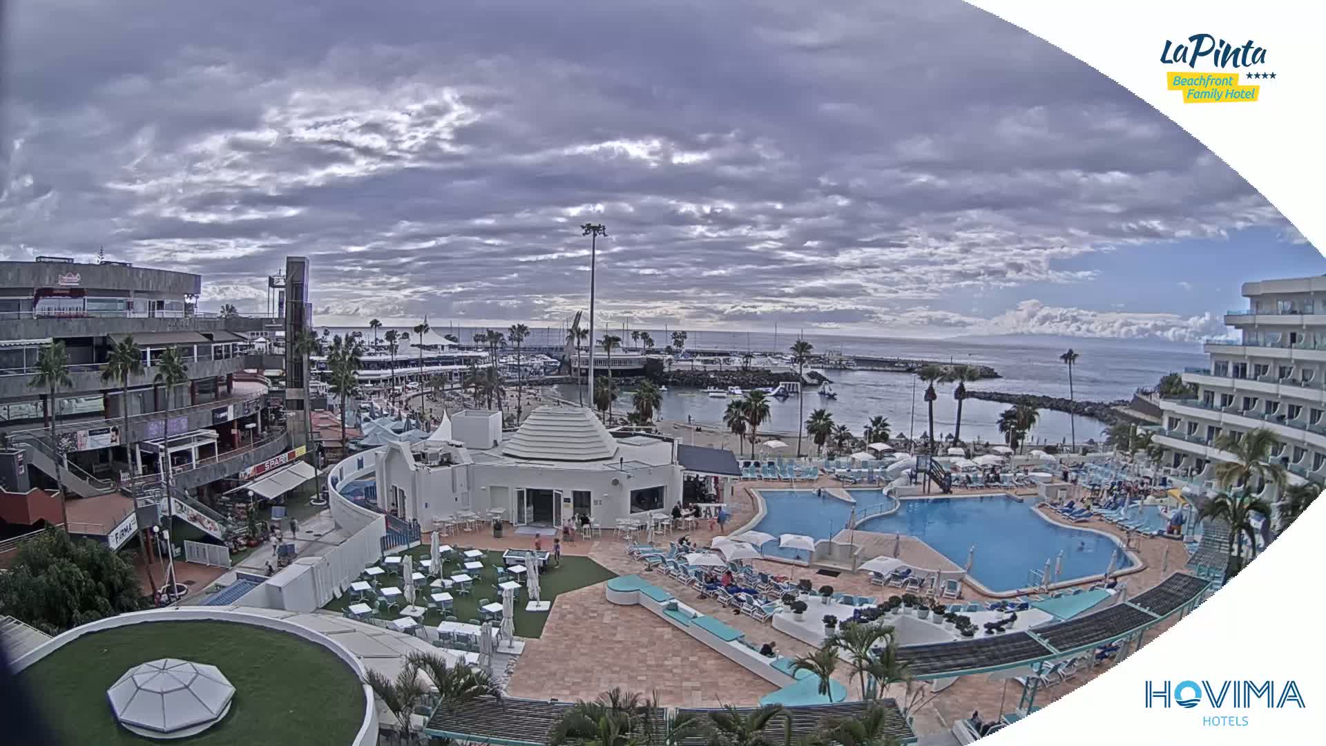 A resort area with a swimming pool and beach, situated near a harbor under a mostly cloudy sky.