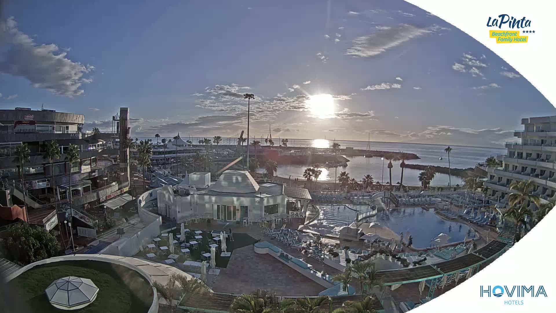A resort with a pool and surrounding buildings overlooks a calm harbor at sunrise under a mostly clear sky.