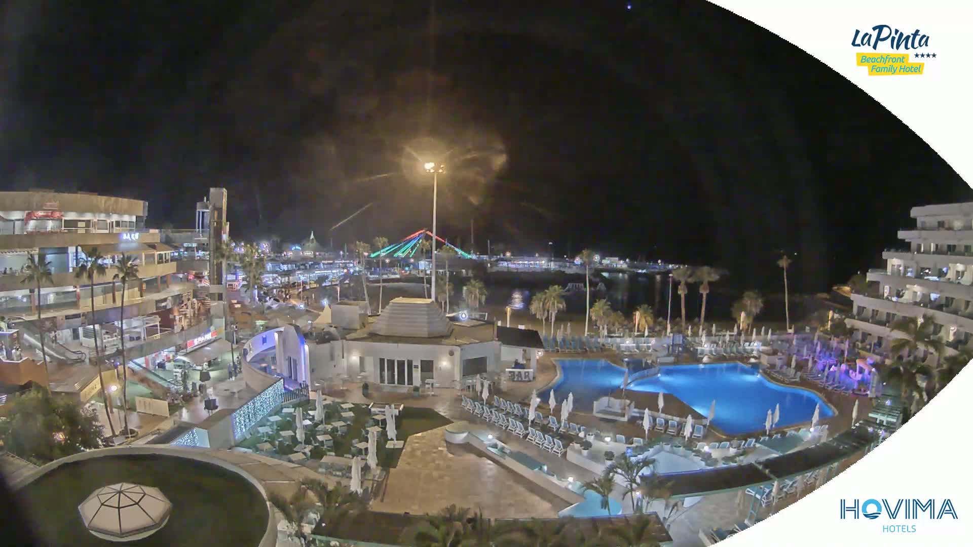 A nighttime aerial view of a resort complex with swimming pools, surrounded by buildings and palm trees, overlooks a beach and a bridge illuminated with colored lights.