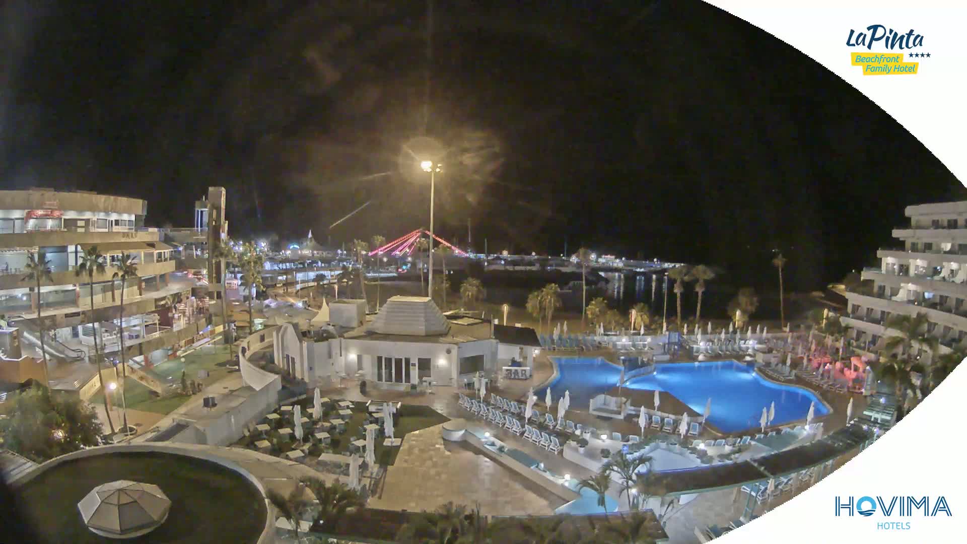 A nighttime aerial view of a resort complex with swimming pools, illuminated buildings, and palm trees, overlooking a calm body of water and a bridge in the distance.