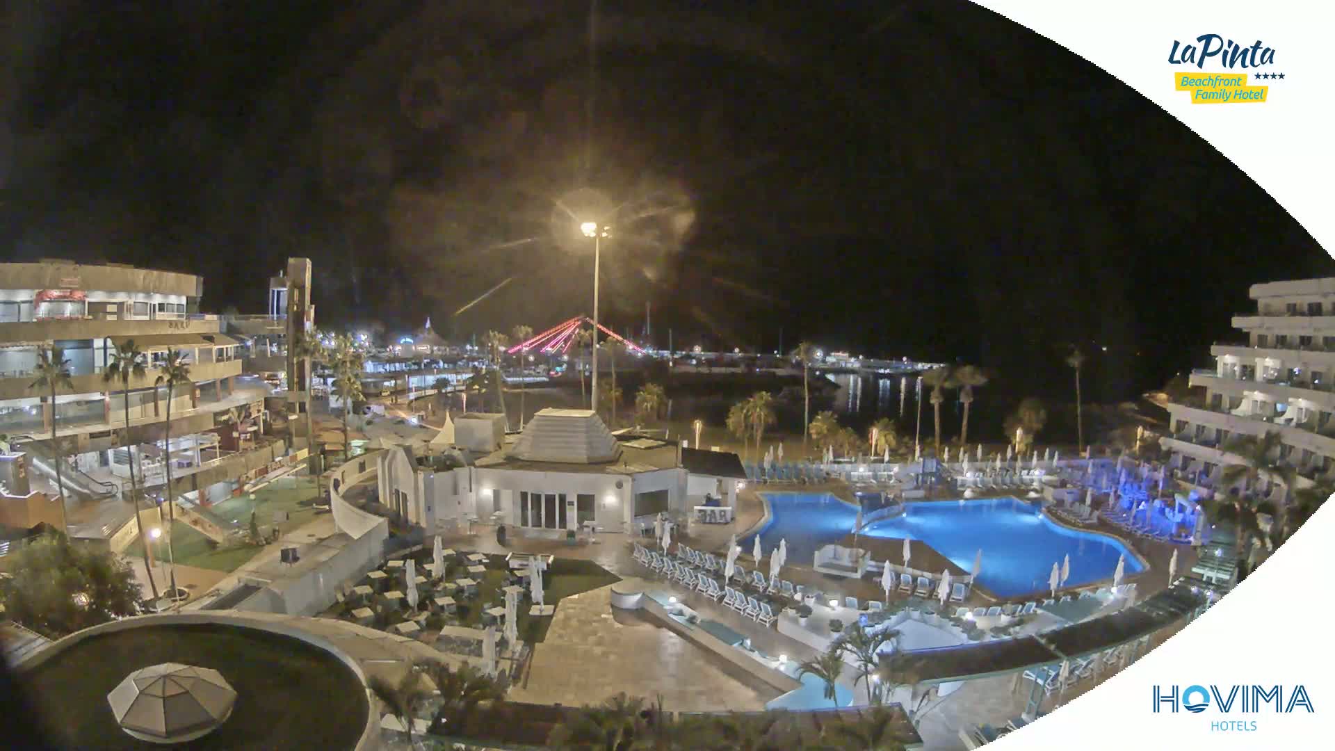 A nighttime aerial view of a resort with swimming pools, surrounding buildings, and a bridge illuminated in the distance.