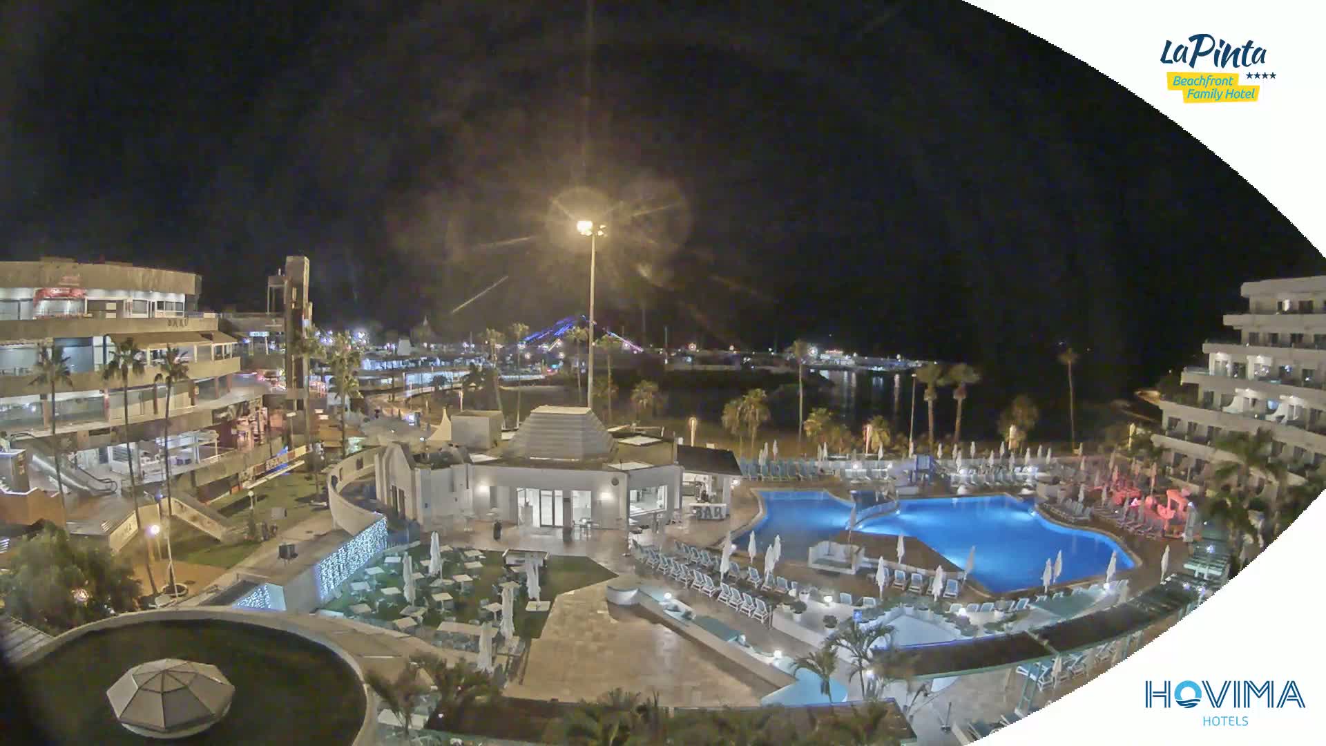 A nighttime aerial view of a resort area with swimming pools, palm trees, and buildings, illuminated under a clear, dark sky.