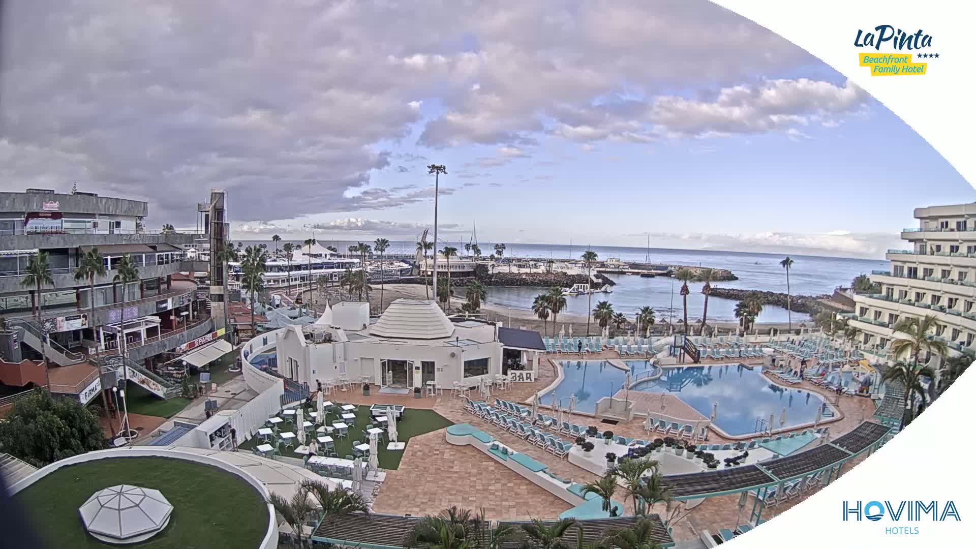 A resort area with a swimming pool overlooks a calm harbor and ocean under a partly cloudy sky.