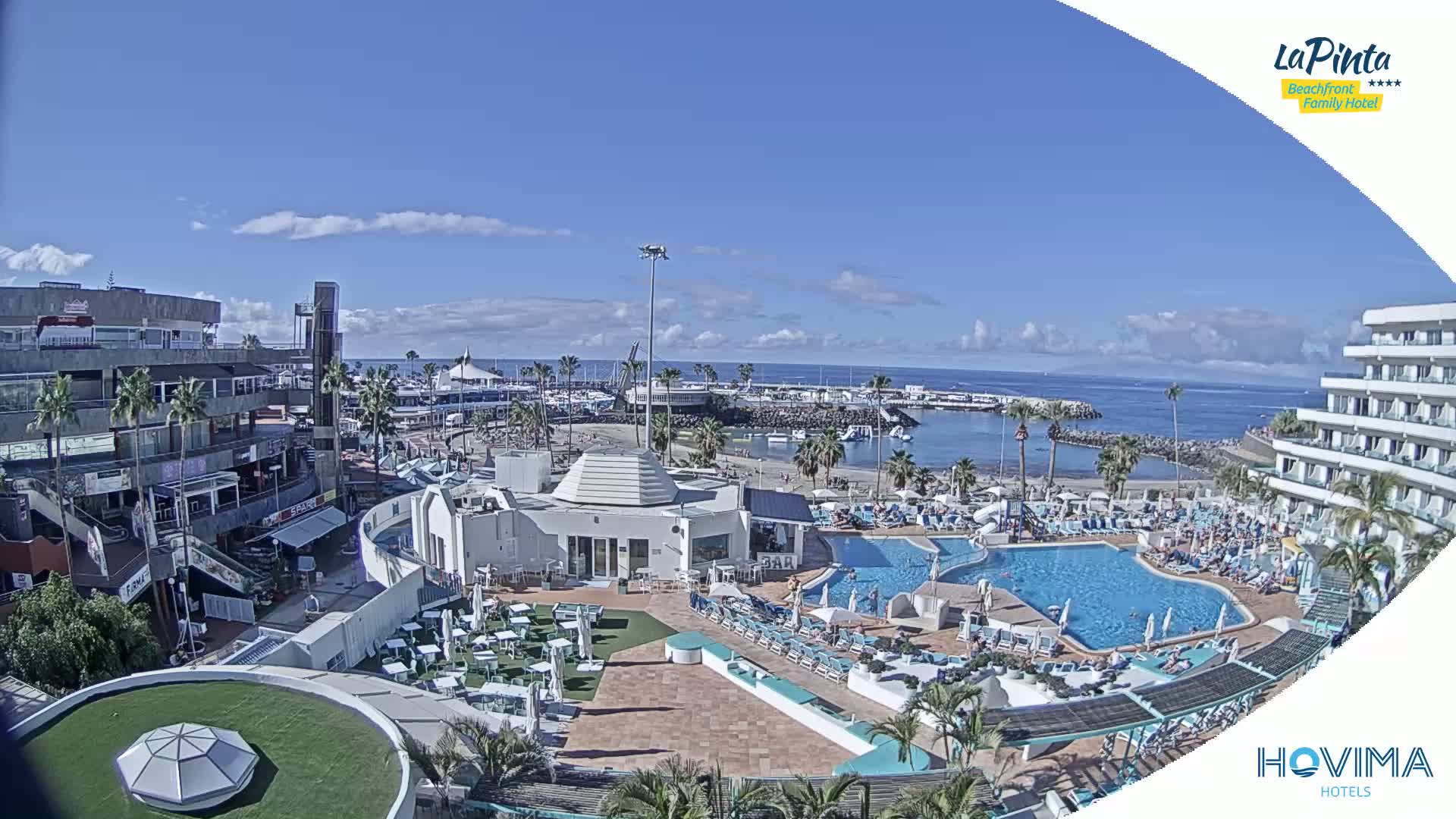 A resort with multiple pools overlooks a marina and ocean under a mostly sunny sky.