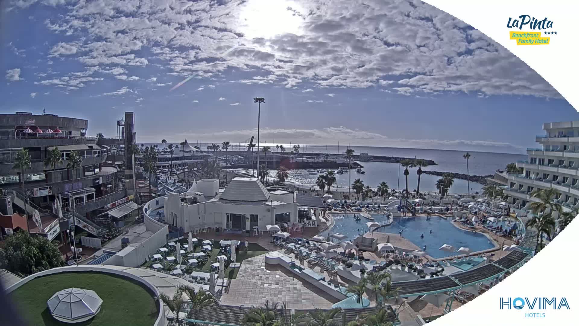 A sunny day overlooking a resort pool area, beach, and harbor, with partly cloudy skies.