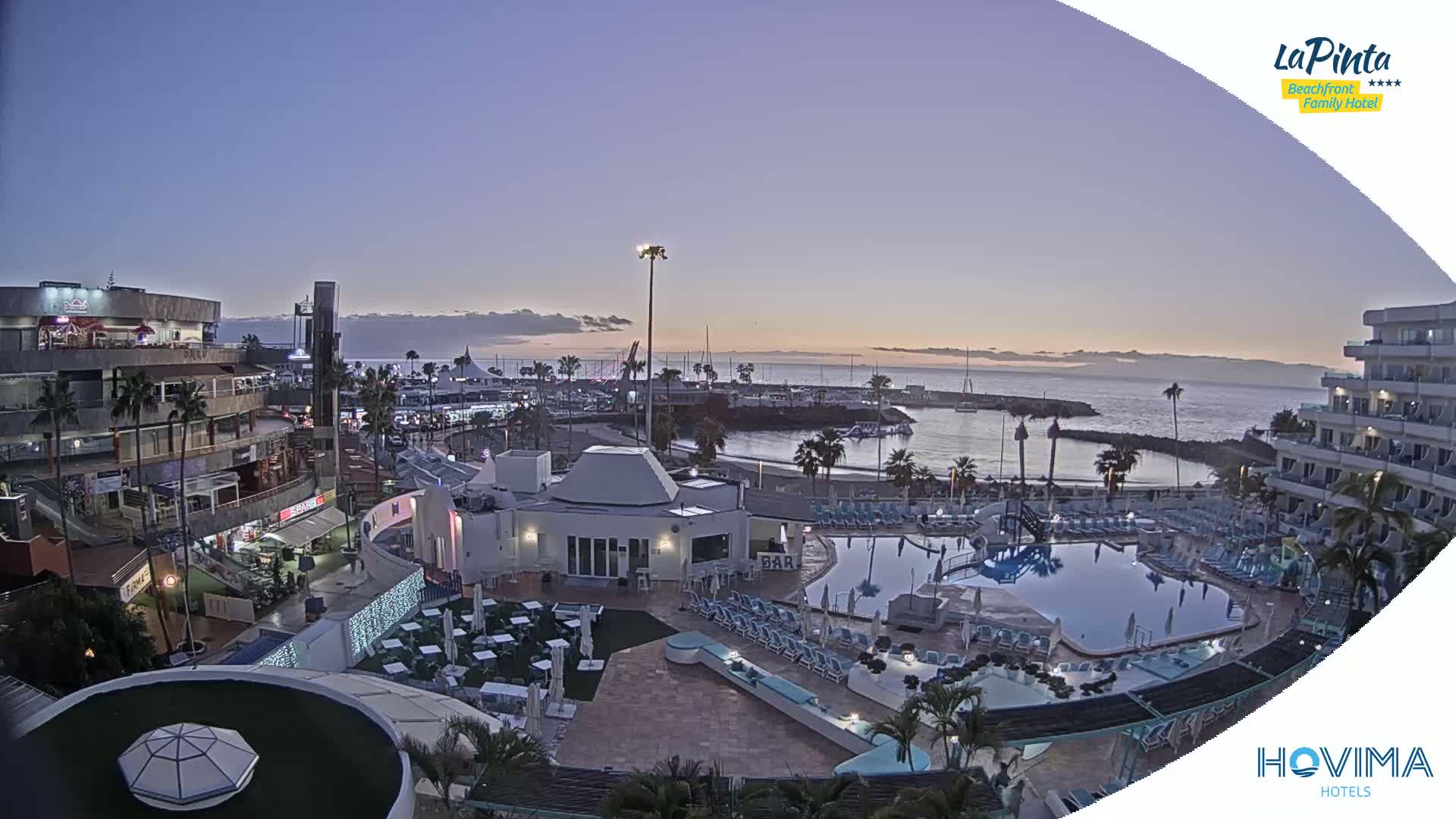 An aerial view shows a resort area at dusk, featuring a swimming pool, buildings, and a harbor with boats under a clear, twilight sky.