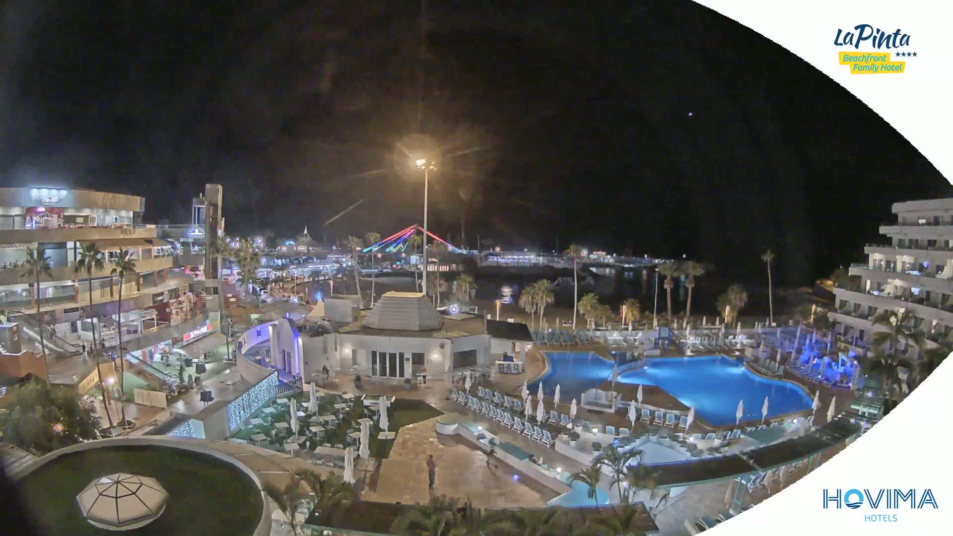 A nighttime aerial view of a resort area with a pool, illuminated buildings, and a bridge in the distance under a clear sky.