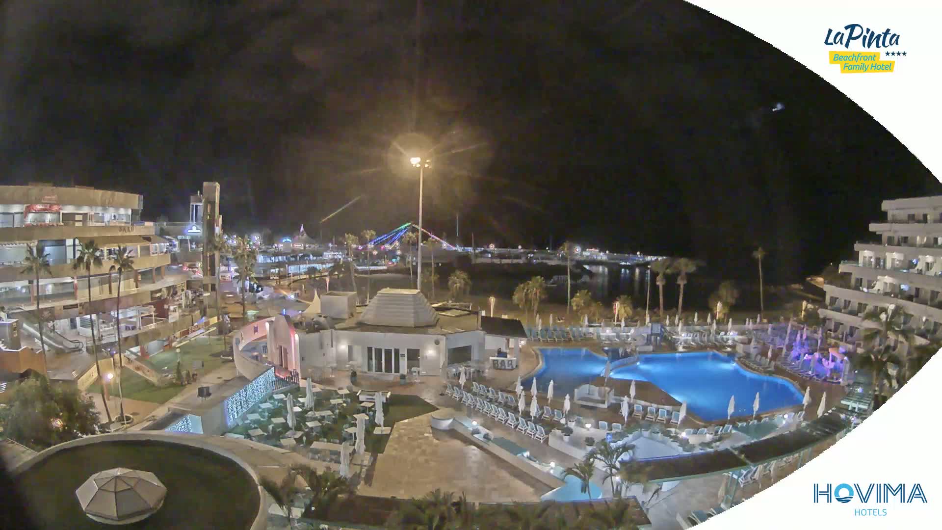 A nighttime aerial view of a resort complex with swimming pools, illuminated buildings, and palm trees, overlooking a calm body of water under a clear, dark sky.