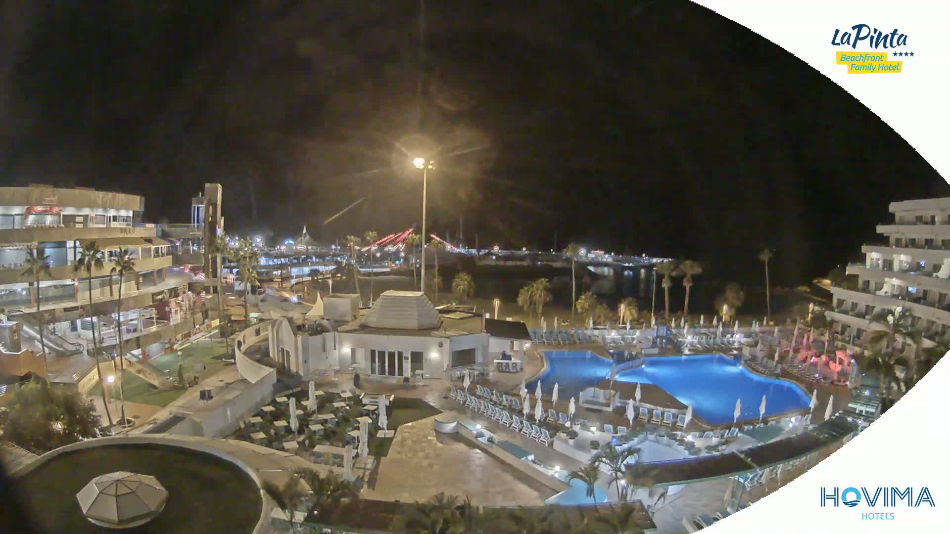 A nighttime view of a resort complex with swimming pools, surrounding buildings, and a beach in the distance under a clear, dark sky.