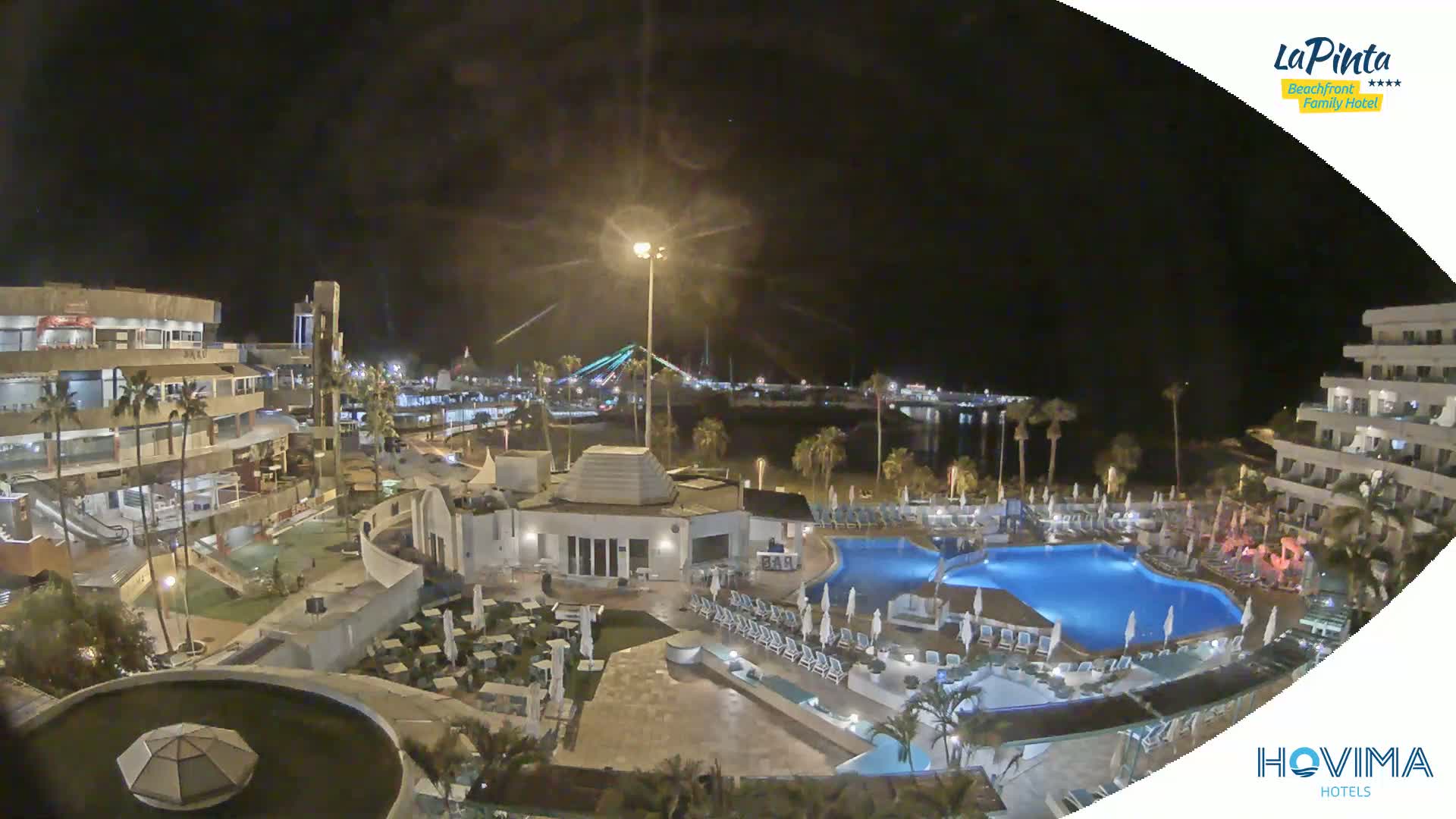 A nighttime aerial view of a resort complex with swimming pools, buildings, and palm trees, overlooking a calm body of water and a bridge in the distance.