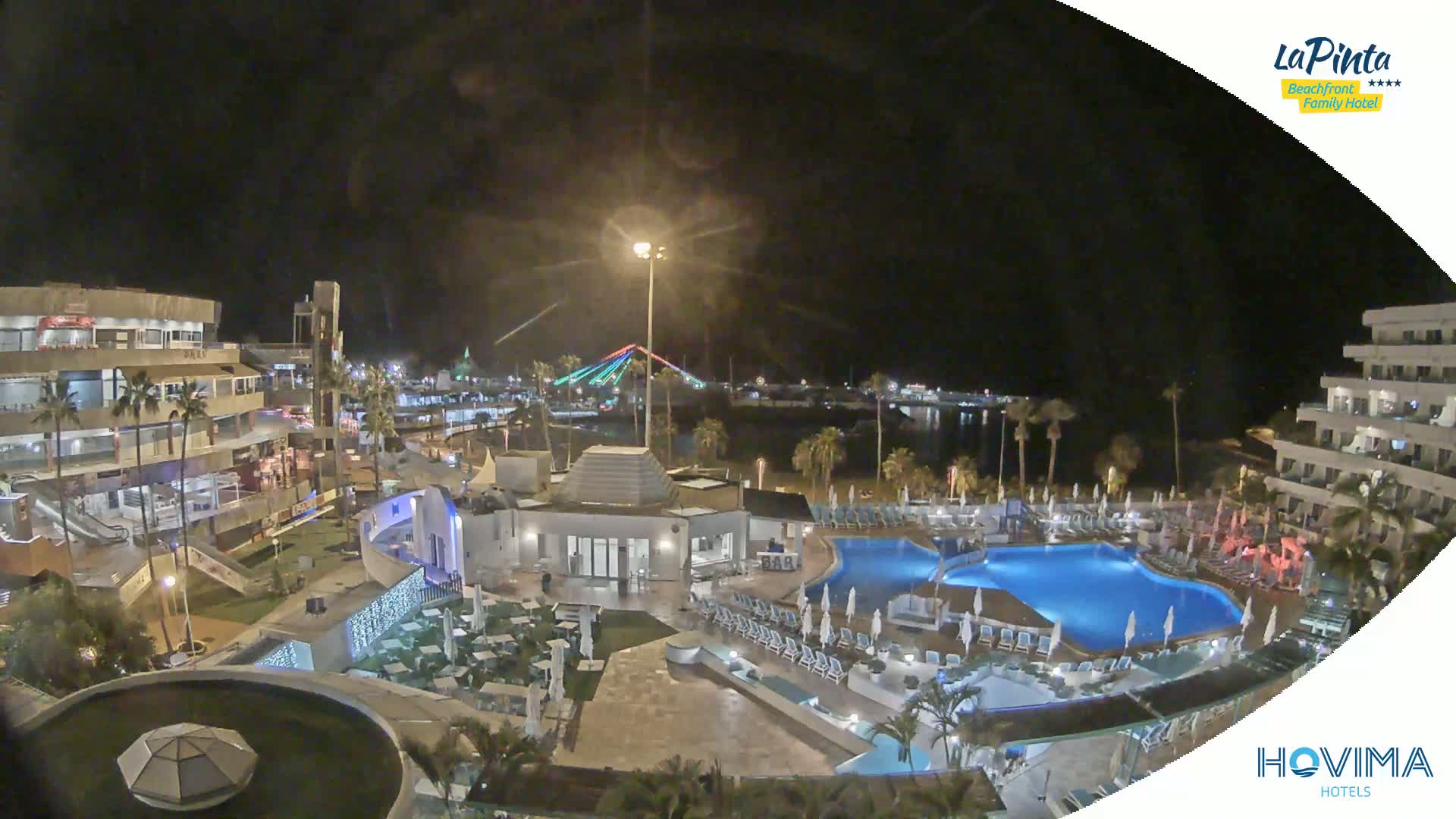 A nighttime view of a resort complex with swimming pools and palm trees, overlooking a calm body of water and a brightly lit bridge in the distance.