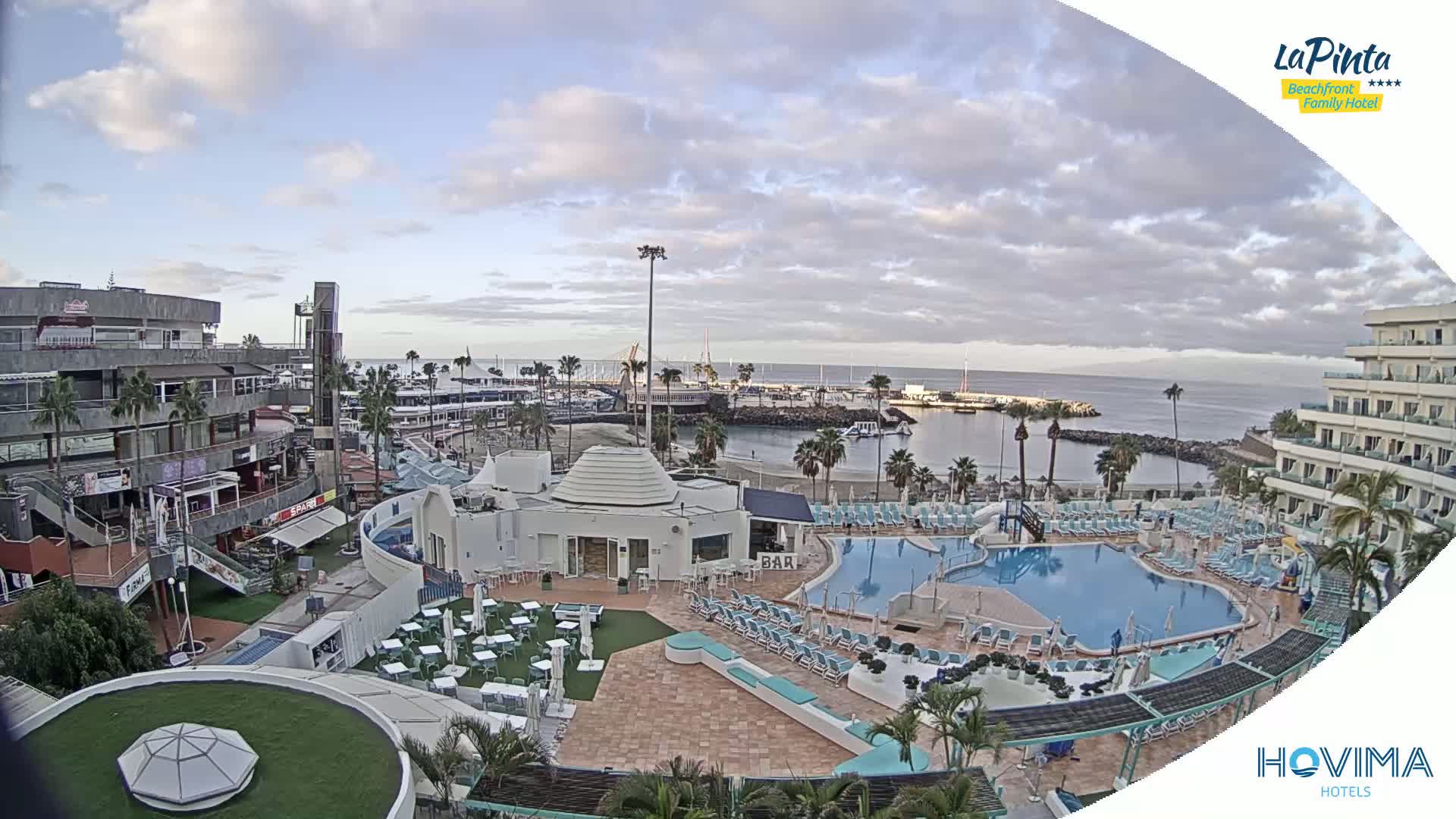 An aerial view of a resort area with a swimming pool, beach, and marina under a partly cloudy sky.