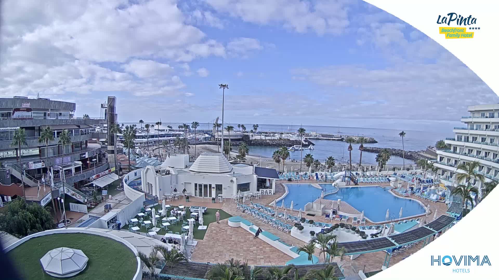 An aerial view of a resort complex with multiple pools, a beach, and a marina, under a partly cloudy sky.