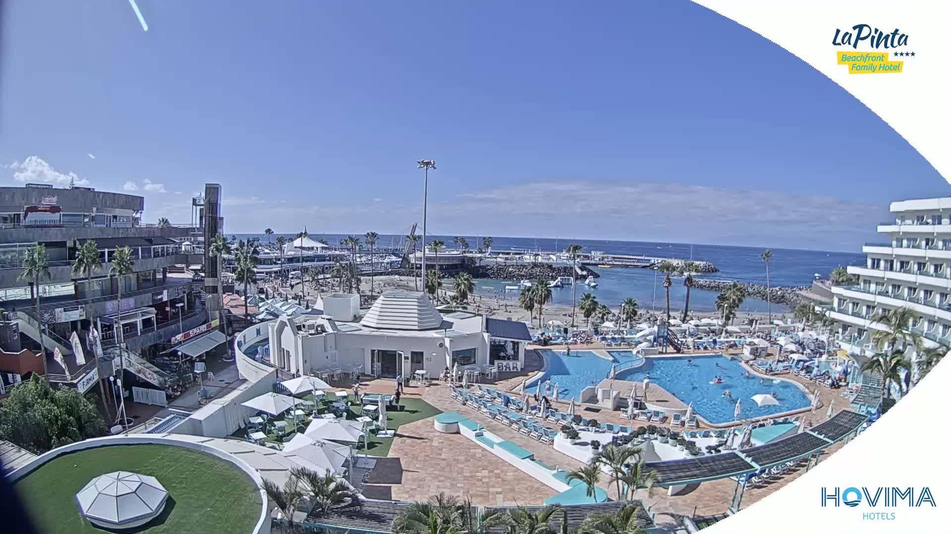 An aerial view shows a resort complex with swimming pools and a beach, all under a clear, sunny sky.