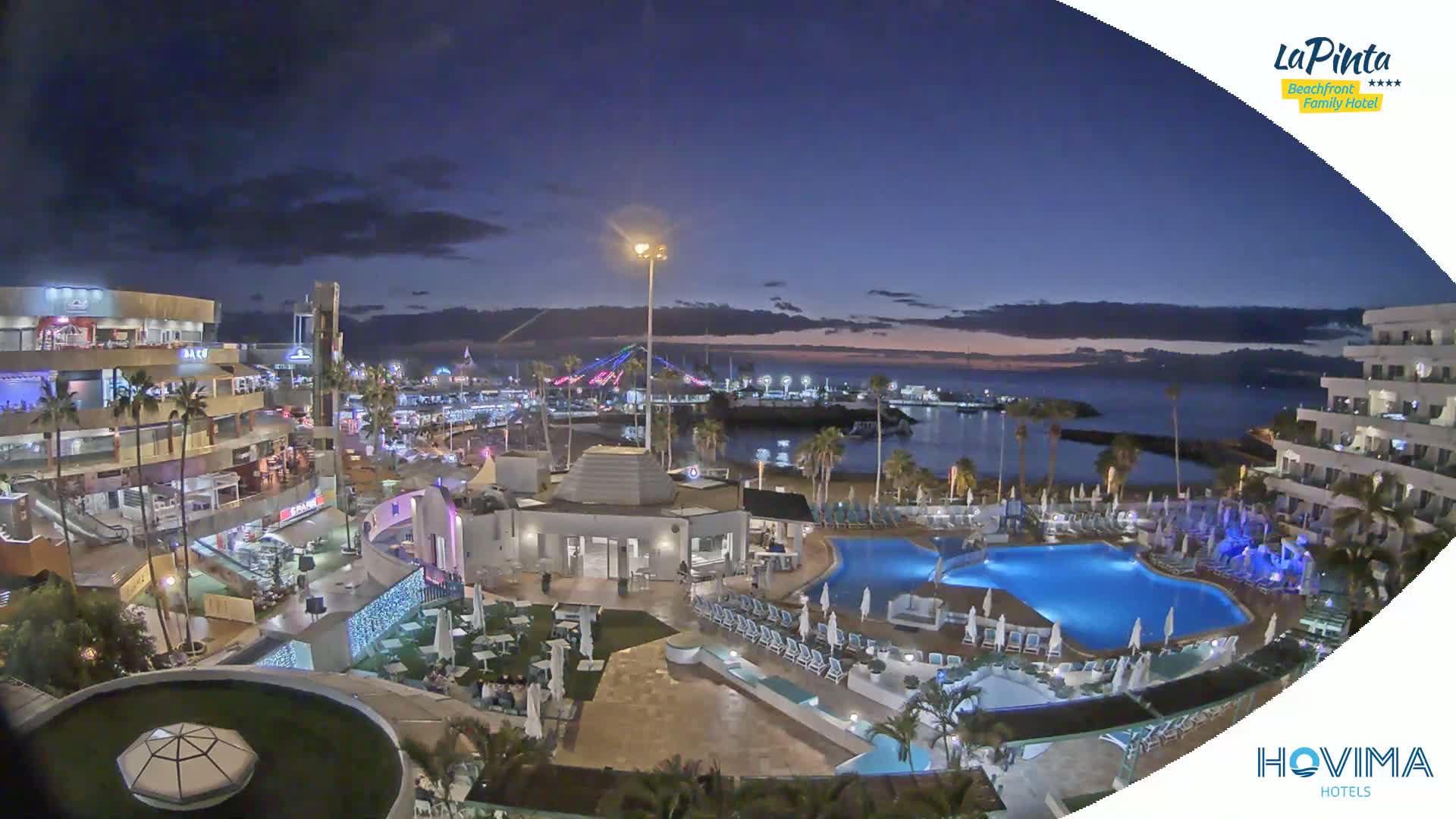 An illuminated resort complex overlooks a calm, dark ocean at twilight, with pools and buildings brightly lit against the darkening sky.