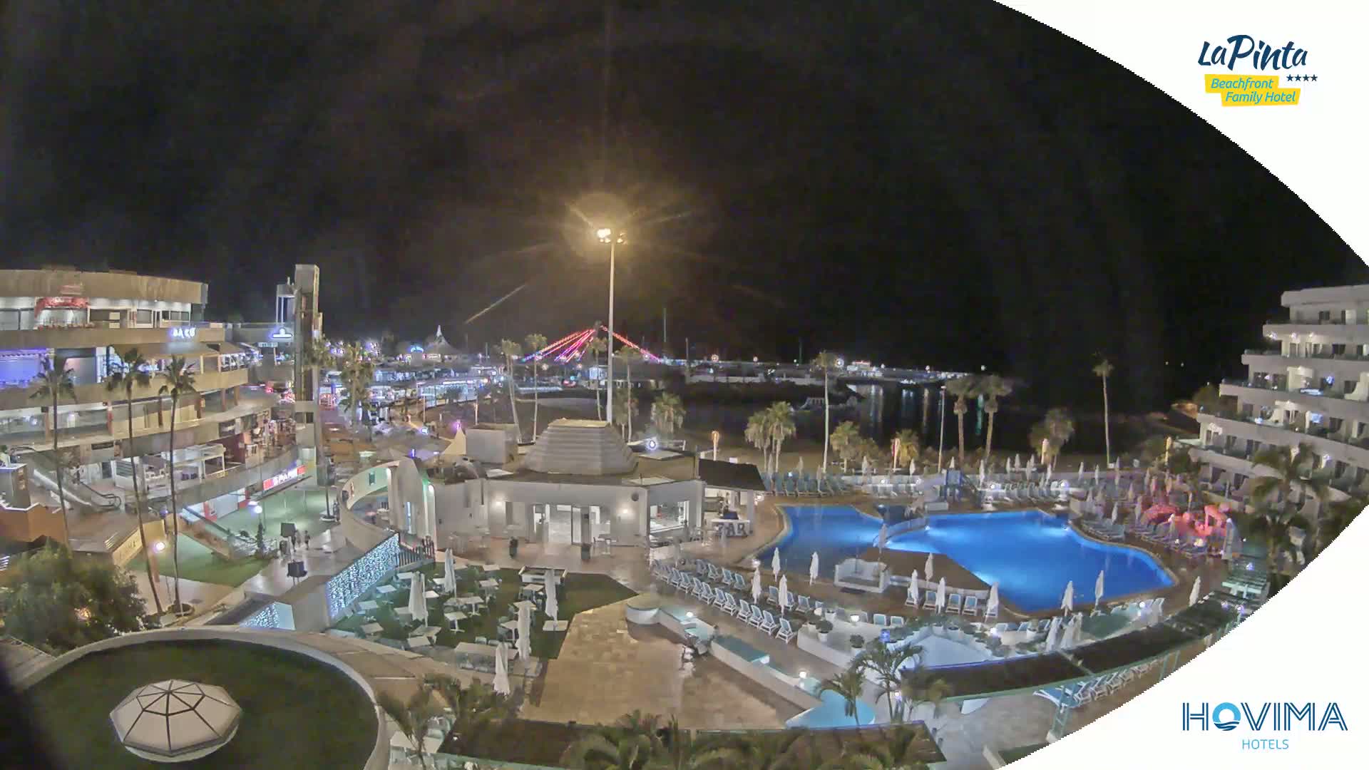 A nighttime aerial view of a resort complex with swimming pools, outdoor seating areas, and nearby buildings, under a clear, dark sky.
