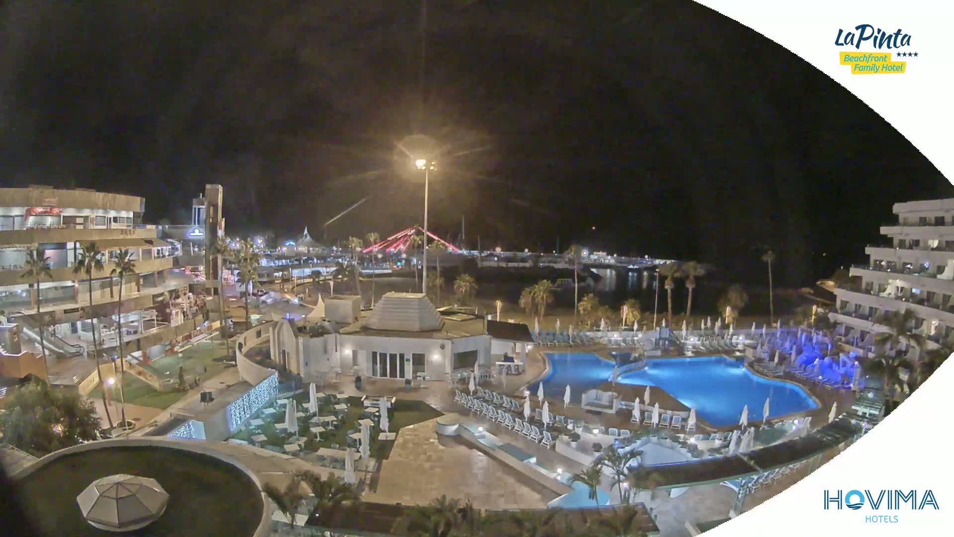 A nighttime aerial view of a resort with illuminated pools and buildings, overlooking a calm body of water and a bridge in the distance.
