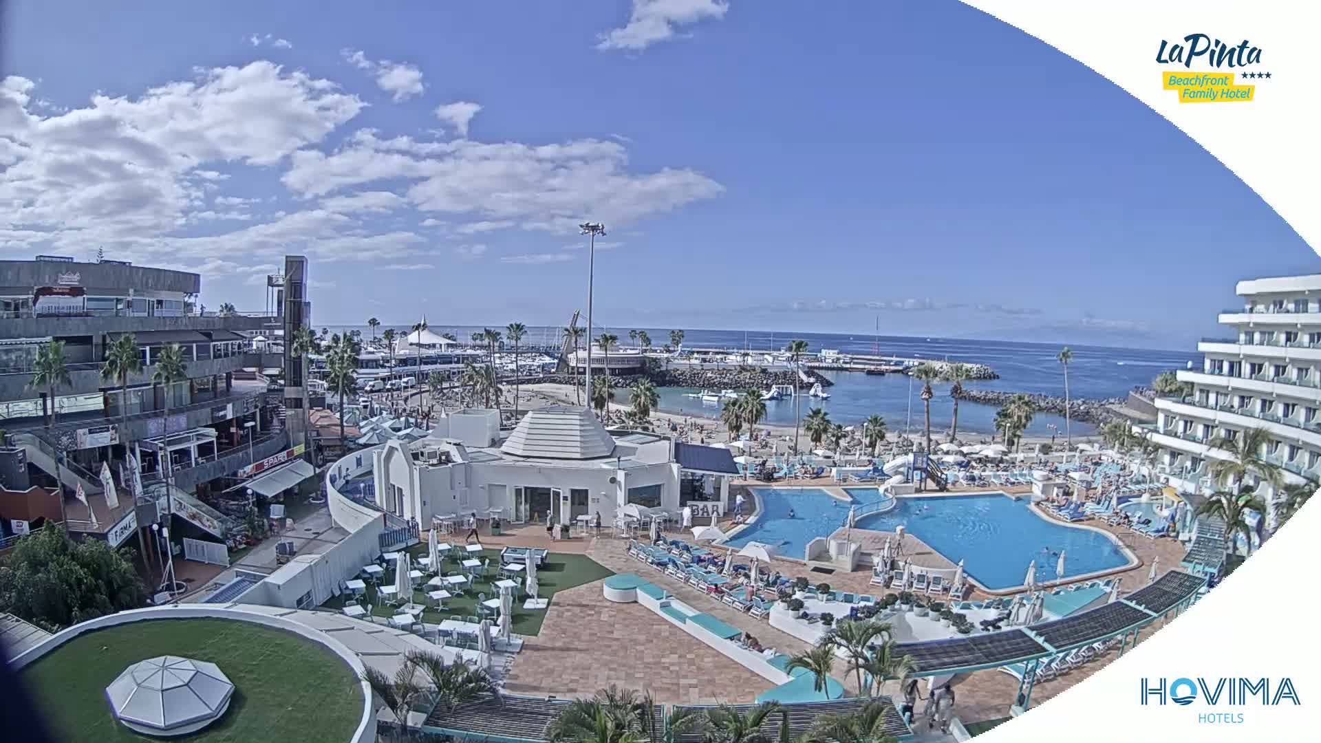 A sunny day at a resort overlooking a calm ocean, featuring a pool area, beach, and surrounding buildings.