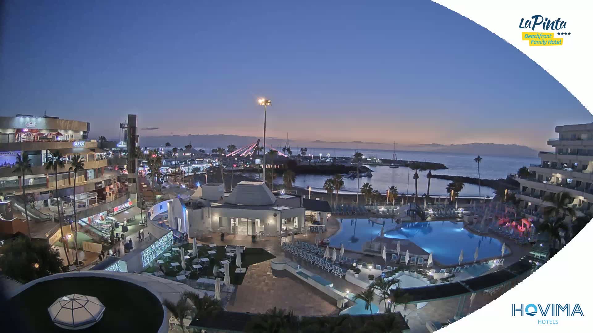 An illuminated resort at twilight overlooks a calm ocean harbor, with palm trees and buildings surrounding a swimming pool.