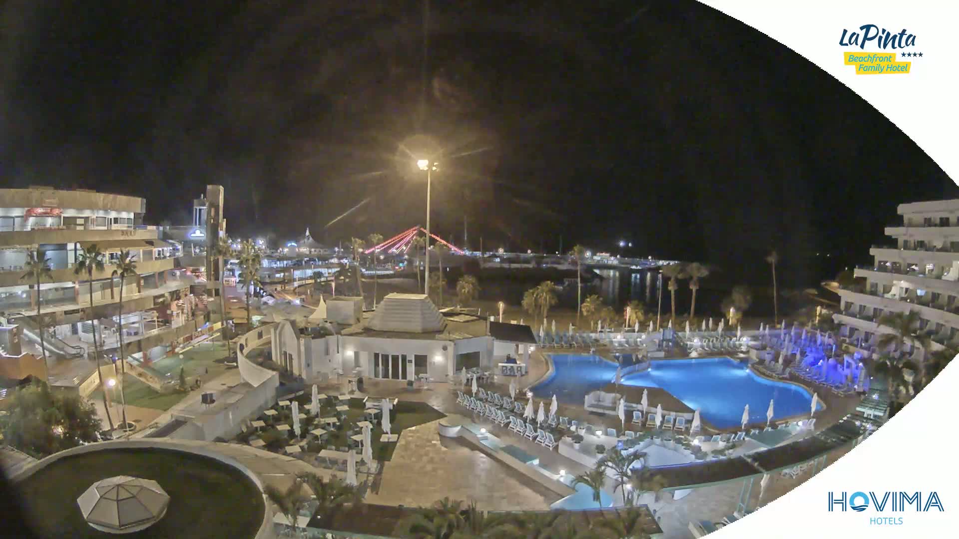 A nighttime aerial view of a resort complex with a swimming pool and surrounding buildings, under a clear, dark sky.