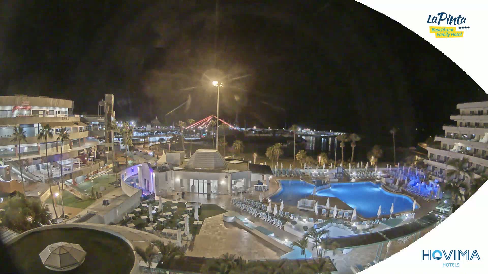 A nighttime aerial view of a resort complex with multiple pools, surrounding buildings, and a waterfront with a bridge in the distance under a clear sky.