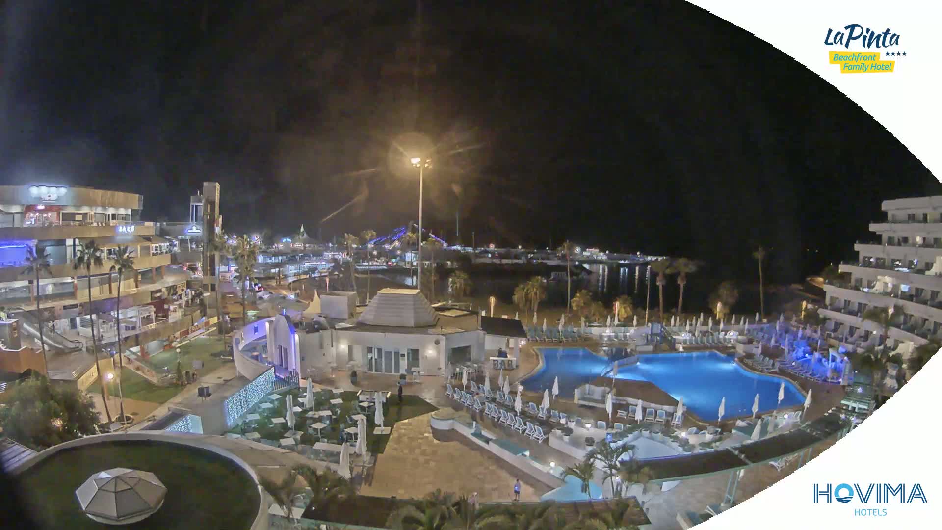 A nighttime aerial view of a resort complex with pools and surrounding buildings, illuminated under a clear, dark sky.