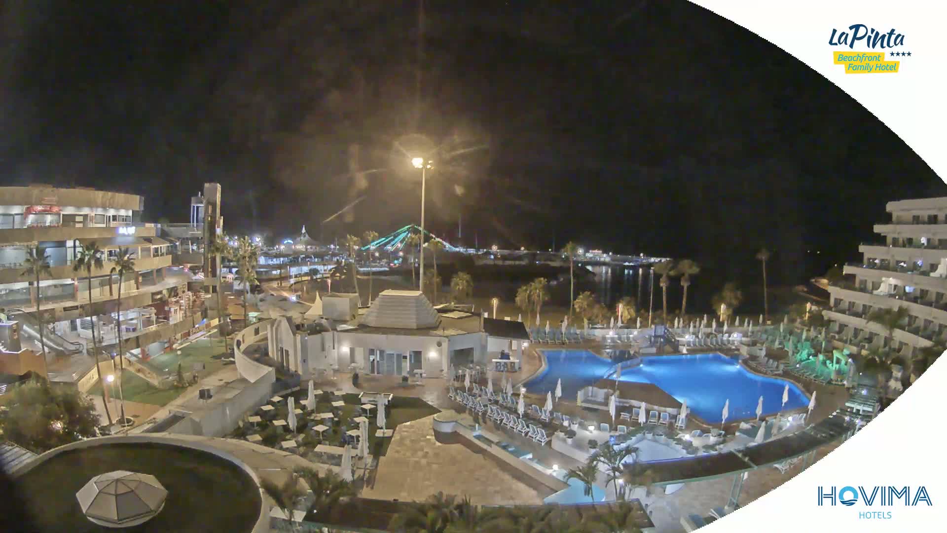 A nighttime aerial view of a resort complex with swimming pools, surrounding buildings, and a beach area illuminated under a clear, dark sky.