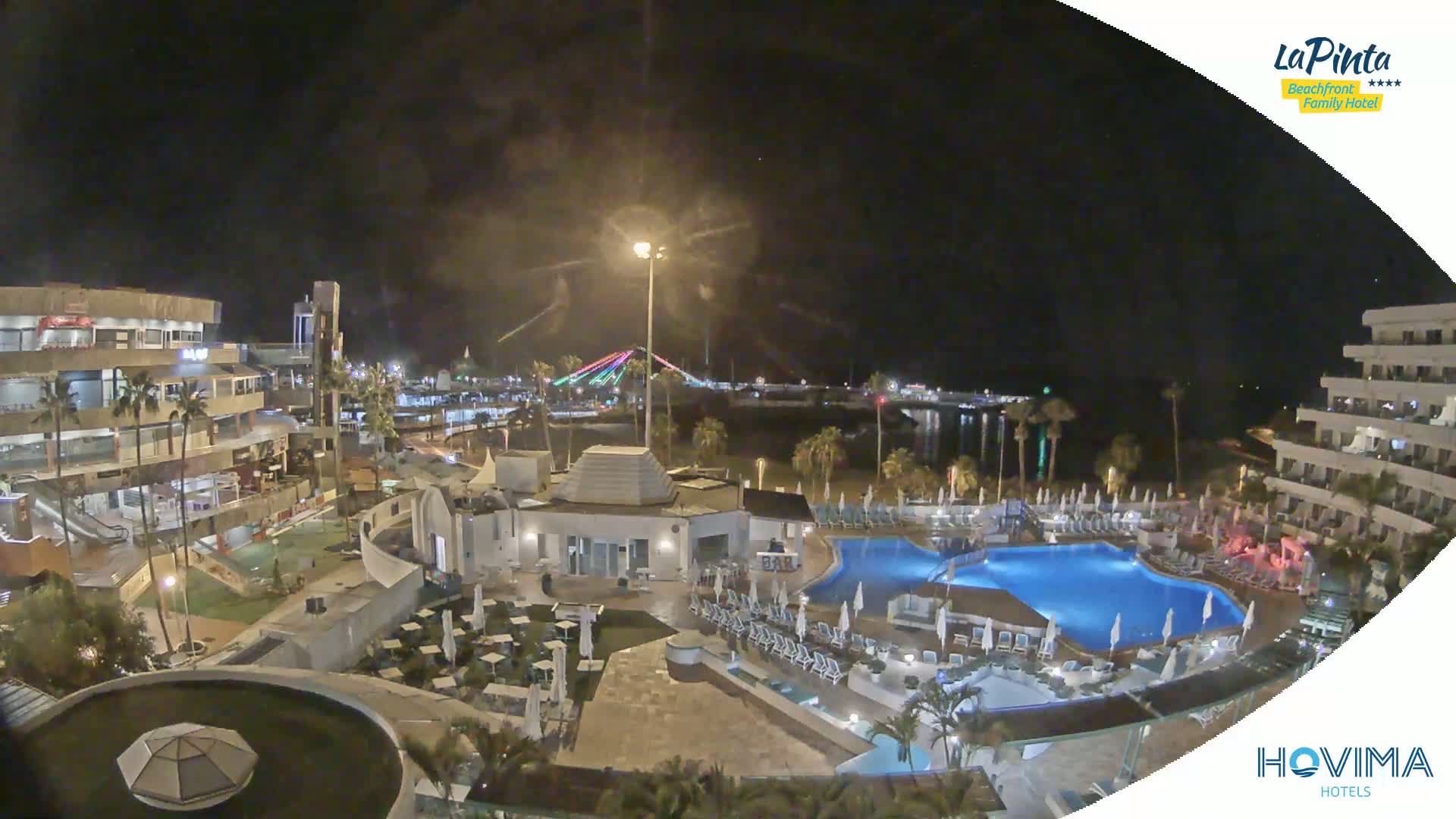A nighttime aerial view of a resort complex with a swimming pool, situated near a waterfront with a lighted bridge in the distance under a clear, dark sky.