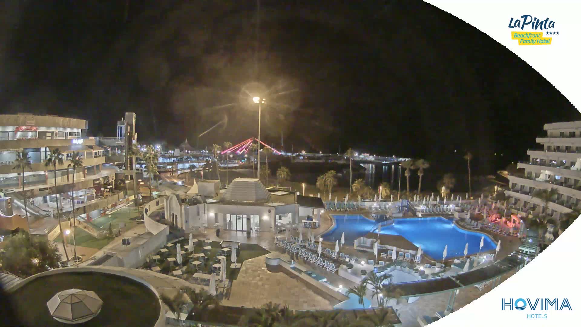 A nighttime view of a resort complex with swimming pools, surrounded by palm trees, near a body of water and a lit bridge.
