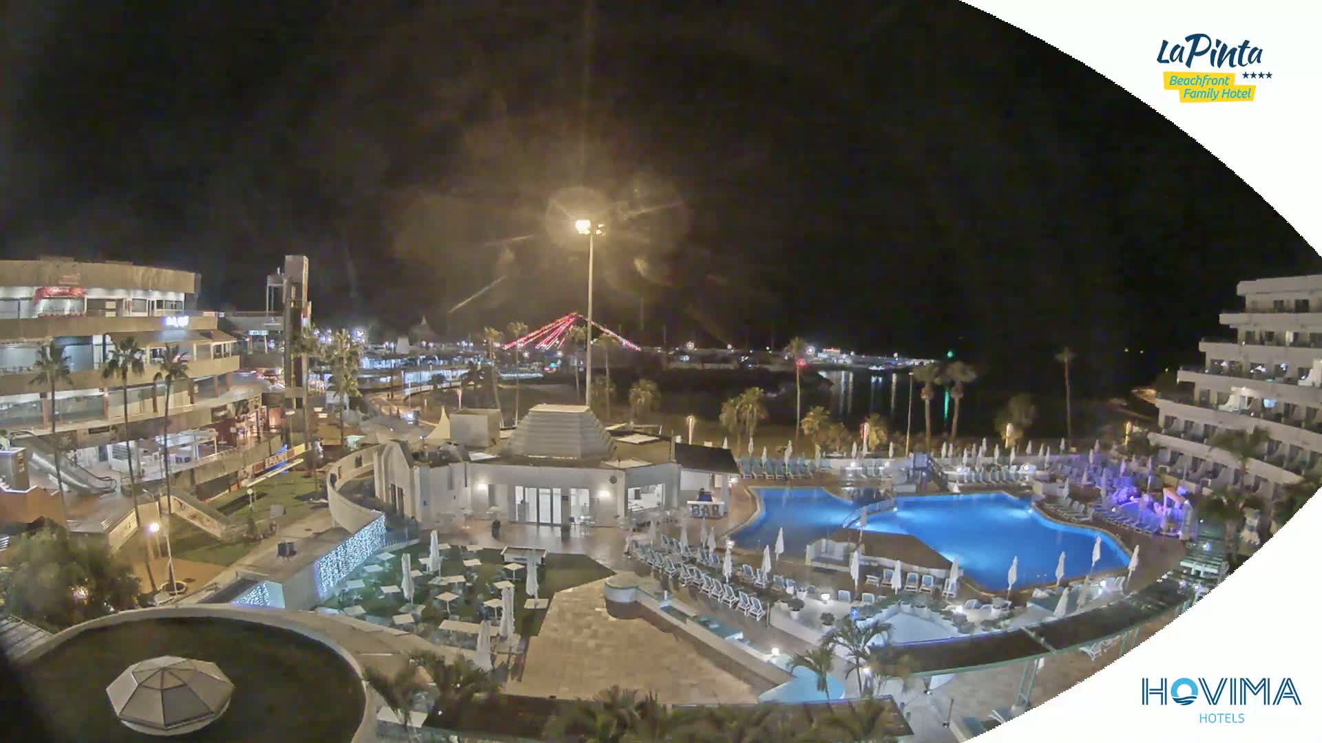 A nighttime aerial view of a resort complex with swimming pools, surrounding buildings, and palm trees, under a dark, cloudy sky.