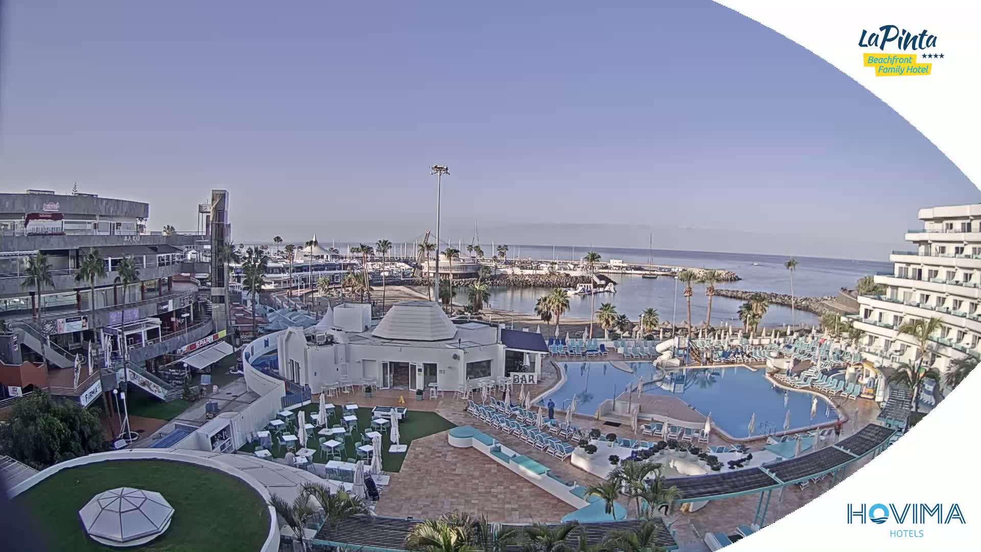 A high-angle view of a resort area with a swimming pool, a marina, and shops under a clear, sunny sky.