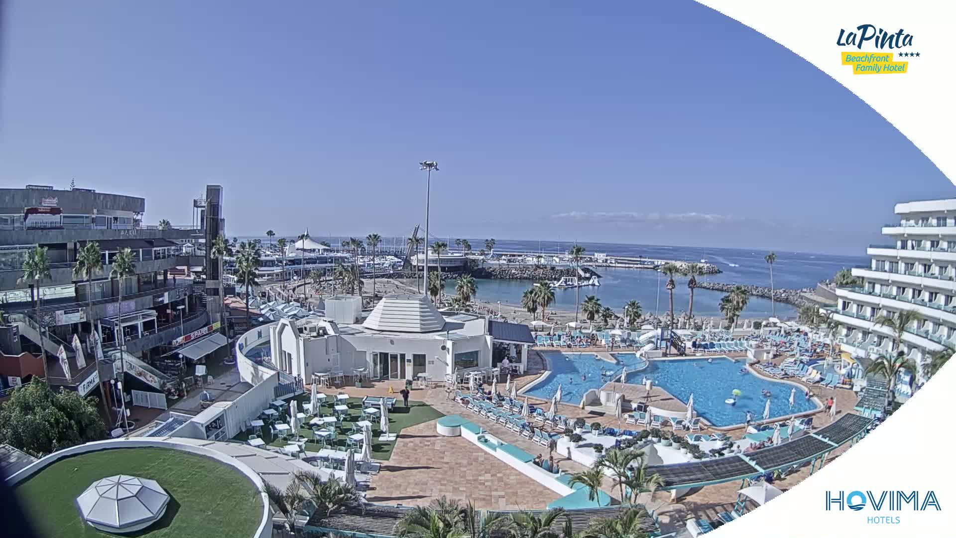 An aerial view shows a resort with a pool and surrounding buildings, overlooking a harbor and calm ocean under a clear, sunny sky.