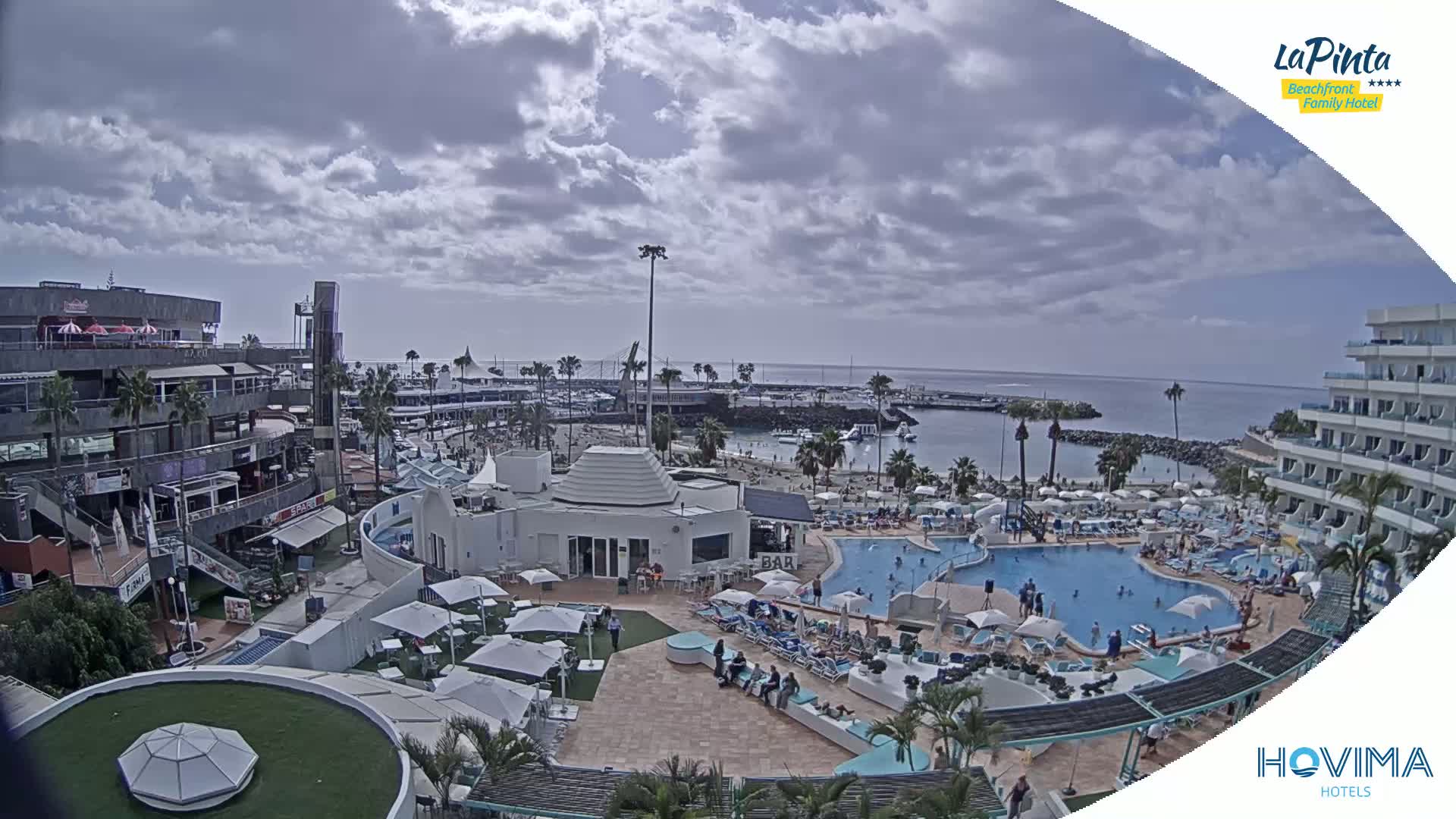 A partly cloudy day overlooks a resort area with multiple pools, a beach, and buildings, including shops and hotels.