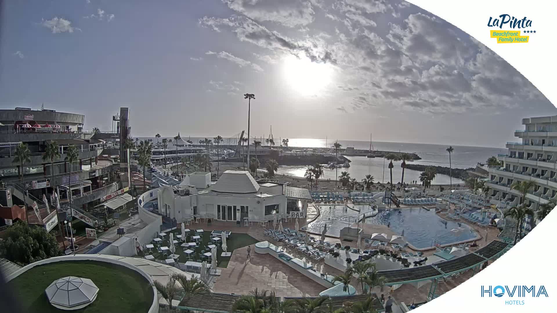 An aerial view shows a resort with a swimming pool and beach area next to a harbor, under a partly cloudy sky with the sun shining brightly.