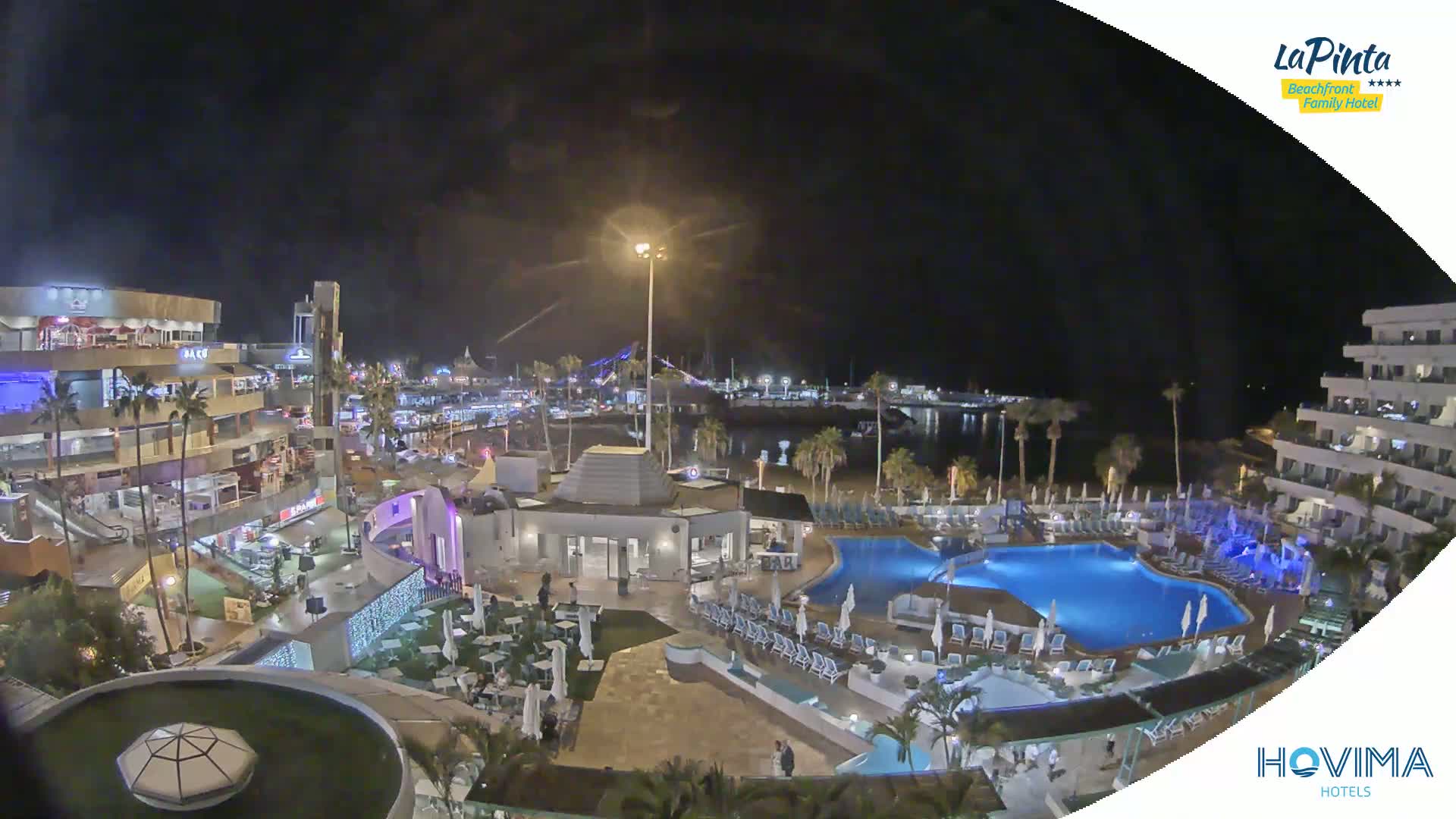 A nighttime aerial view of a resort complex with pools, situated near a waterfront area with palm trees and buildings under a clear, dark sky.