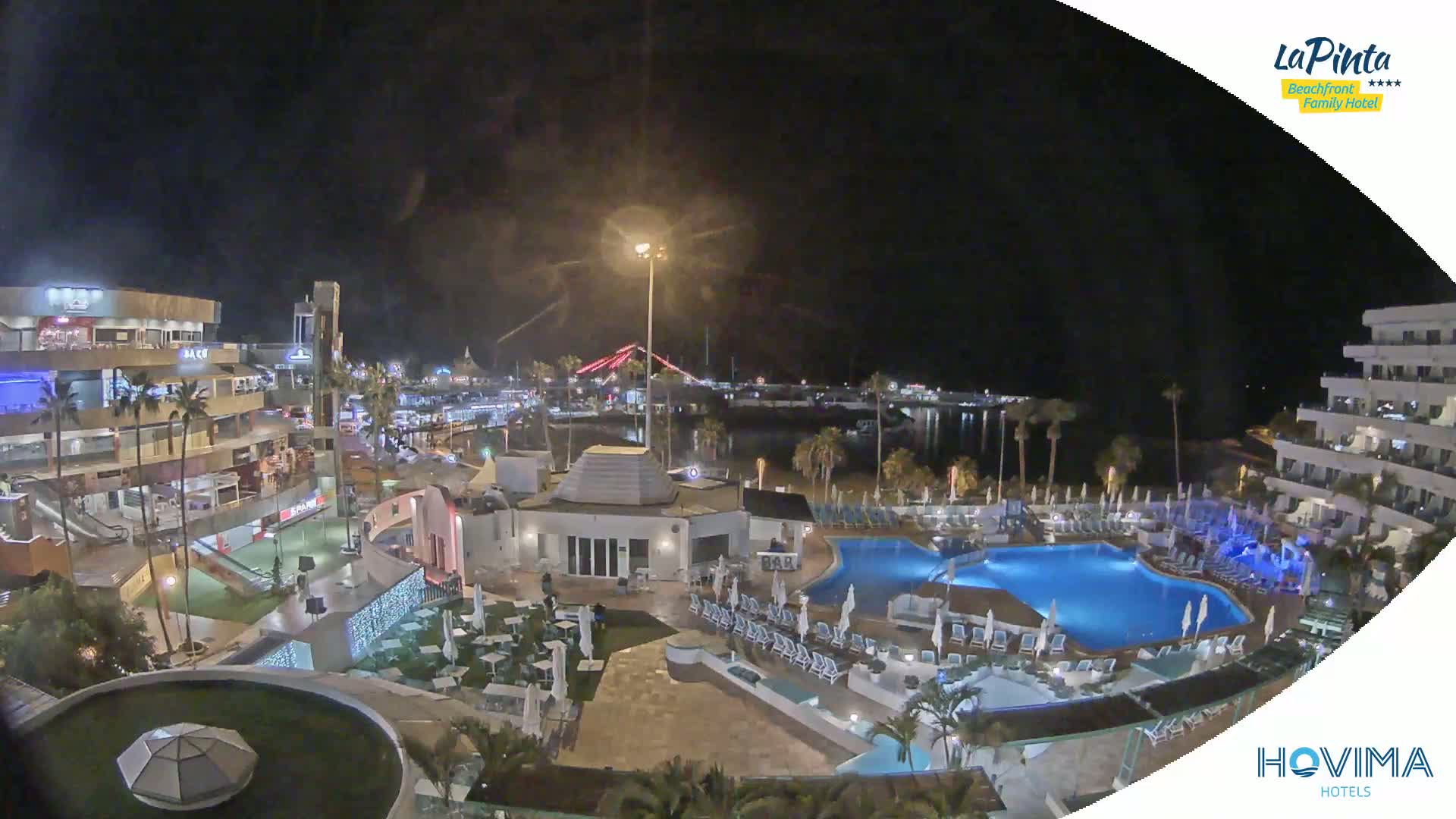 A nighttime aerial view of a resort with swimming pools and surrounding buildings near a waterfront, under a clear, dark sky.