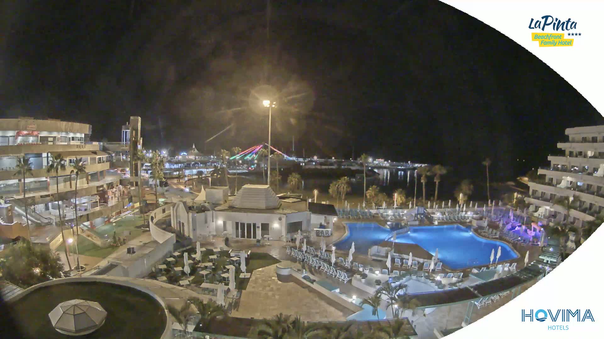 A nighttime view of a resort complex with multiple pools, surrounding buildings, and palm trees, overlooking a calm body of water with a bridge in the distance.