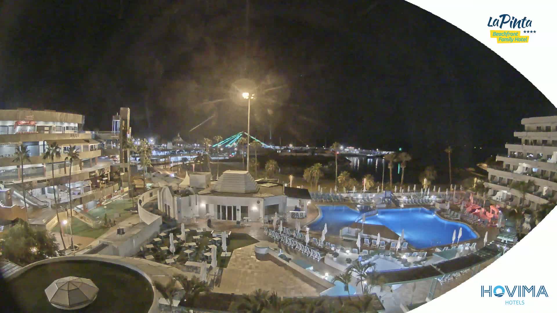A nighttime aerial view of a resort complex with a swimming pool, surrounded by buildings and palm trees, overlooking a body of water and a bridge in the distance.