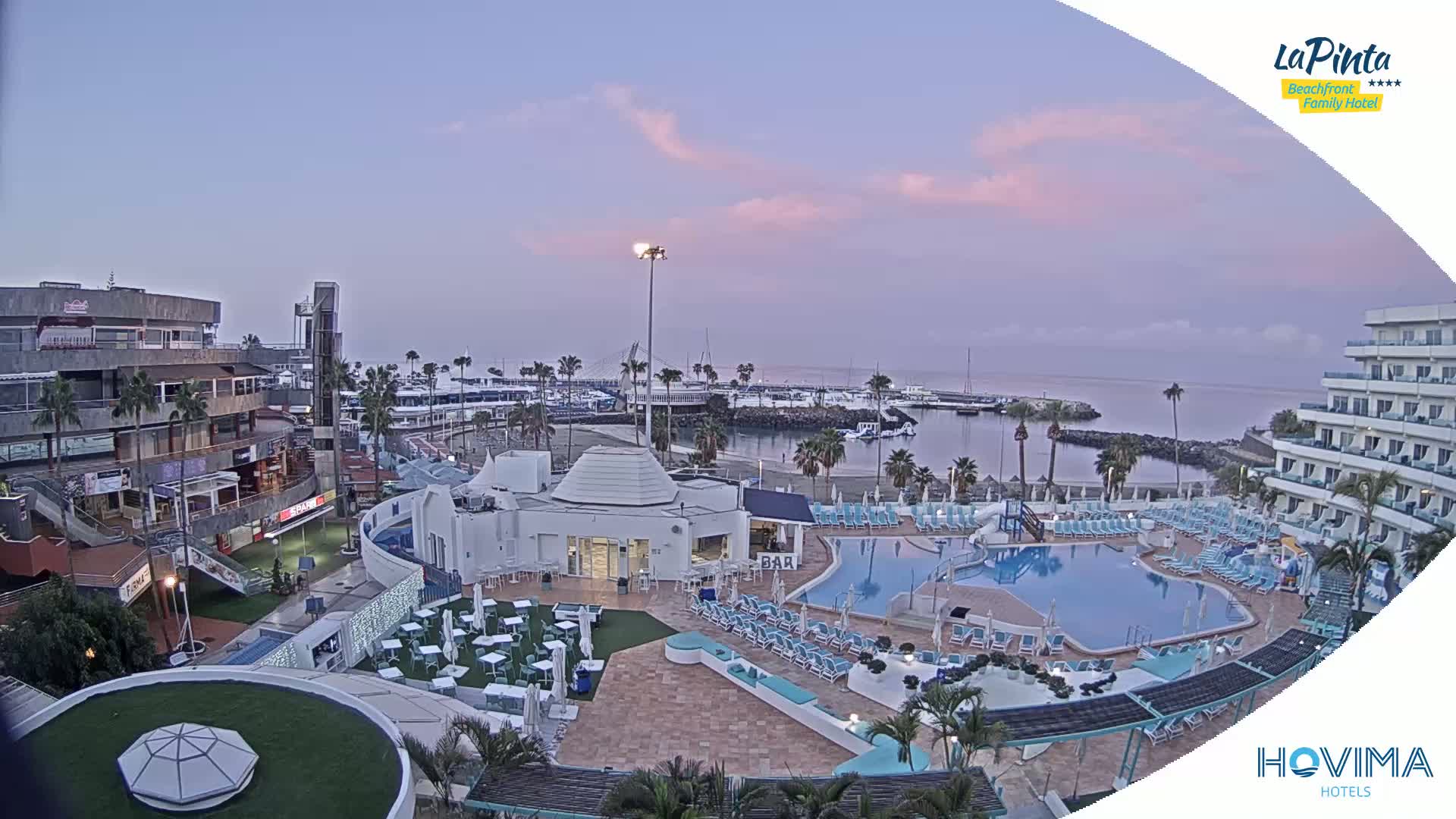 An outdoor view at dawn shows a resort with a swimming pool and surrounding buildings next to a marina, under a pale pink and purple sky.