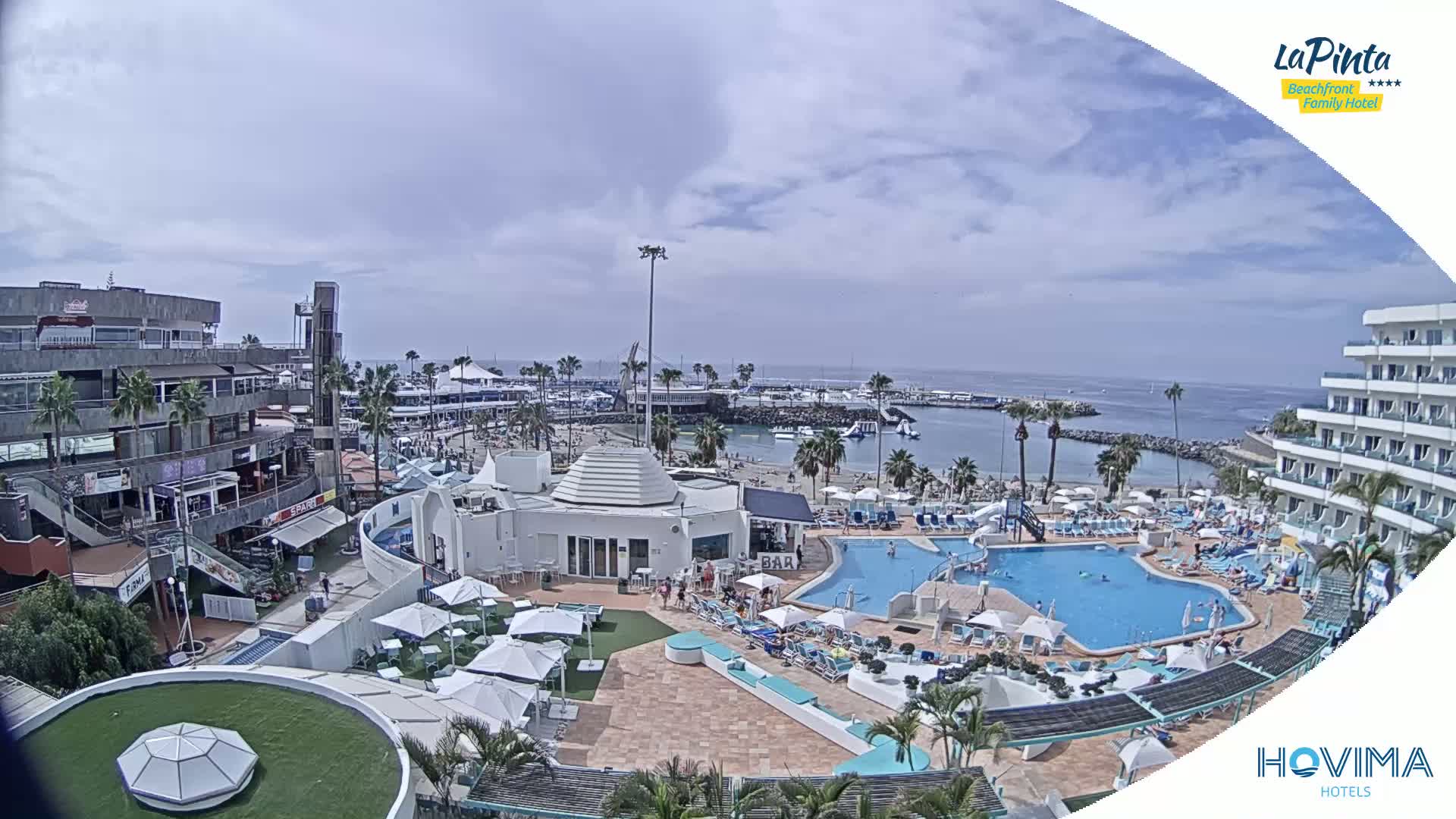 An outdoor scene shows a resort pool area with a beach and calm ocean in the background under partly cloudy skies.