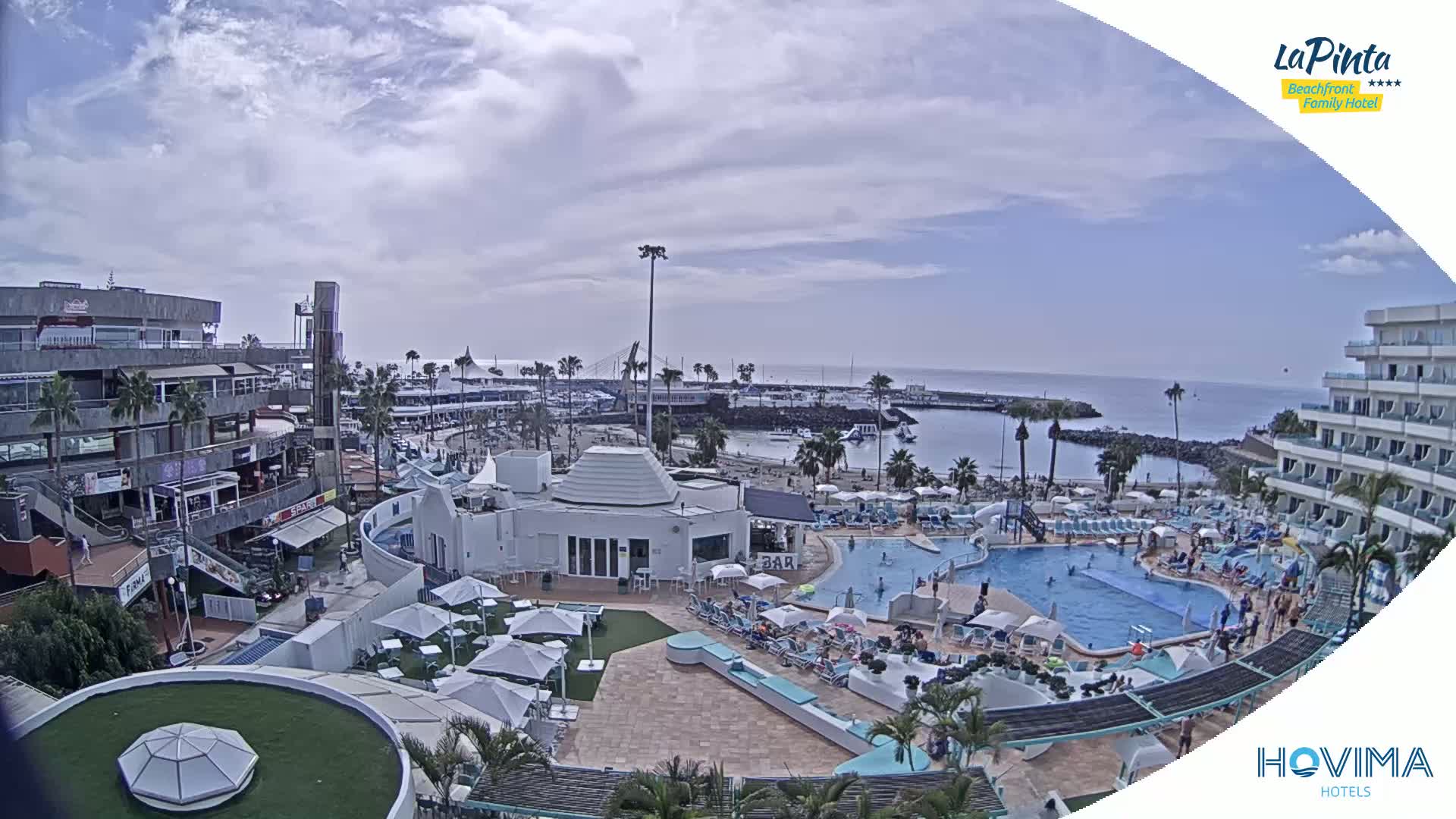 An outdoor scene shows a resort area with a swimming pool, beach, and marina under partly cloudy skies.