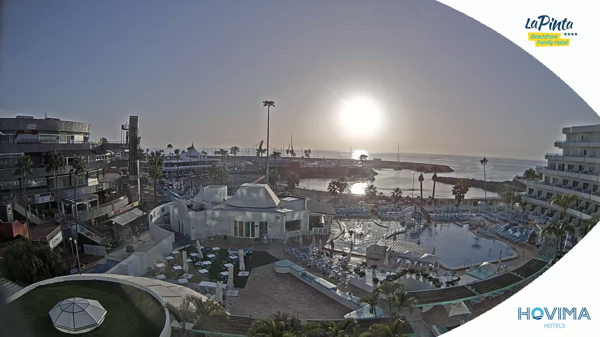 A sunny, outdoor view of a resort with a pool, shops, and a marina overlooking a calm ocean at sunrise.