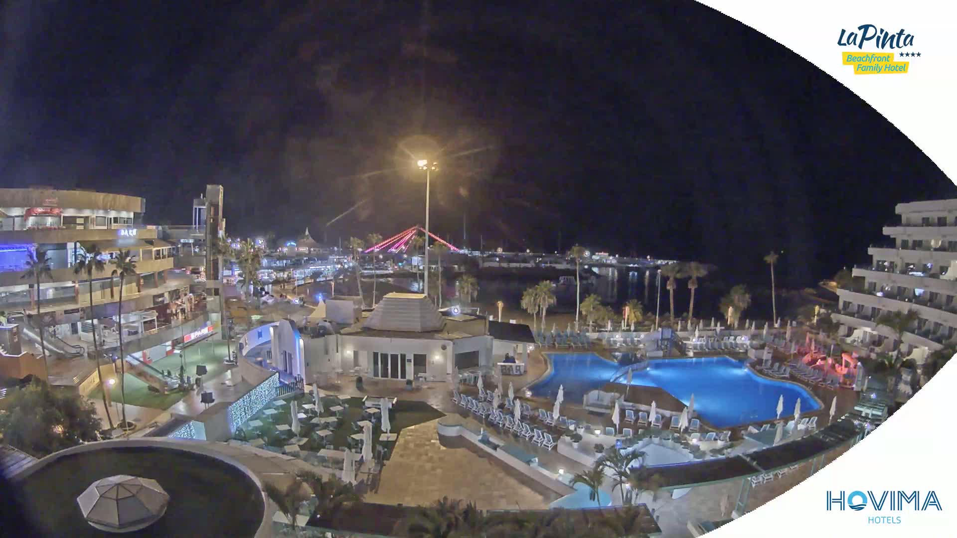 A nighttime aerial view of a resort complex with swimming pools, surrounding buildings, and a waterfront area with a bridge illuminated in the distance.