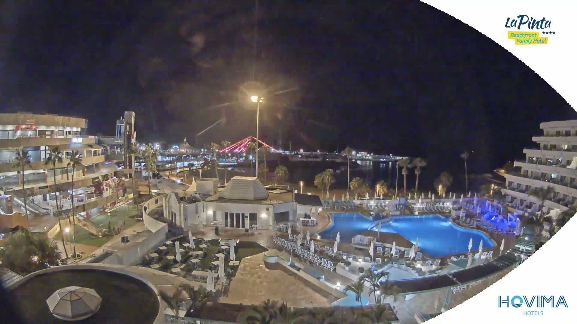 A nighttime aerial view of a resort complex with a swimming pool, surrounding buildings, and a body of water with a bridge in the distance under a clear sky.
